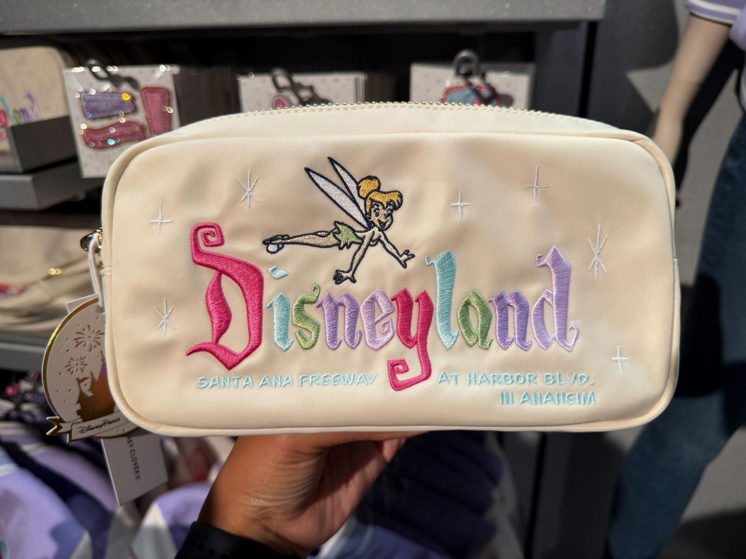 This white pouch by Stoney Clover has a pastel, multi-colored Disneyland embroidery with Tinker Bell flying above it. In light blue embroidery below it is the address information for the park.