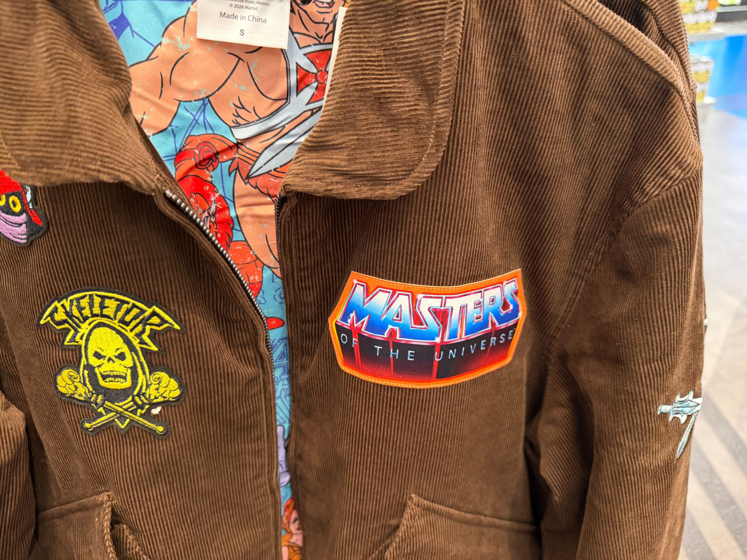 The brown corduroy jacket features "Masters of the Universe," "Skeletor," and "Orko" patches.