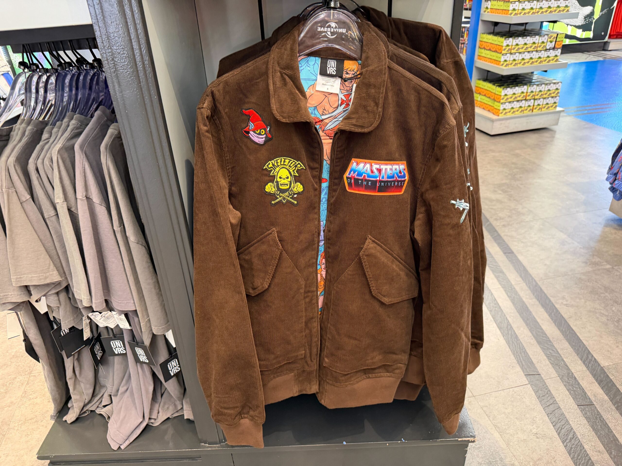 The brown corduroy jacket features "Masters of the Universe," "Skeletor," and "Orko" patches.