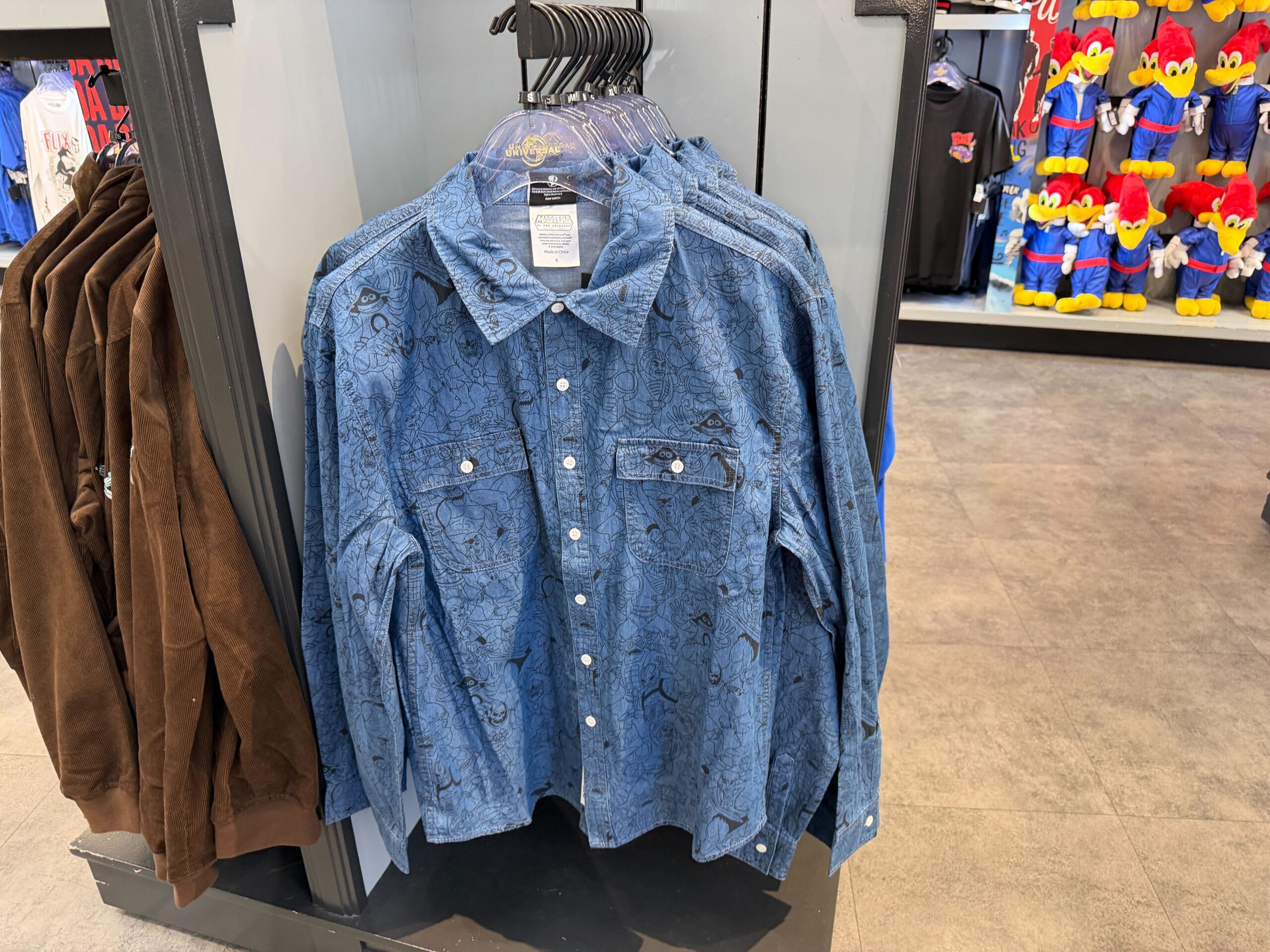 The long-sleeve denim shirt features a collar and two chest pockets. There are white buttons. It has an all-over print of He-Man characters.