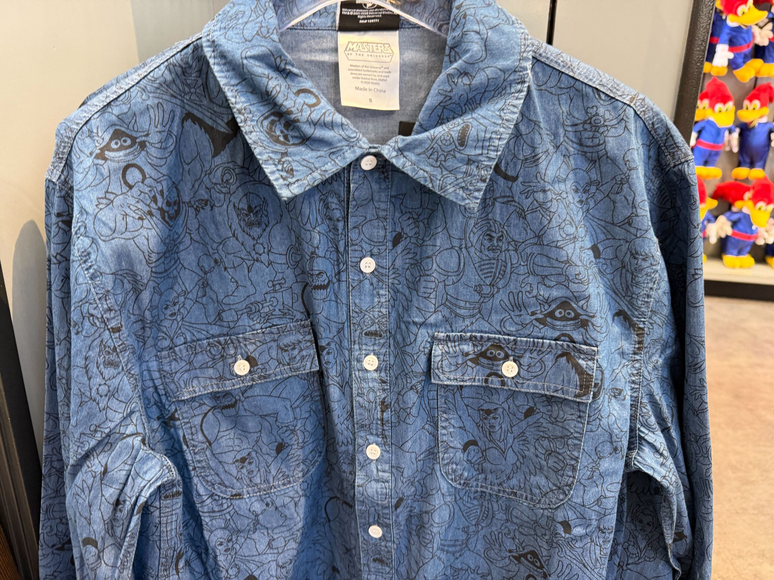 The long-sleeve denim shirt features a collar and two chest pockets. There are white buttons. It has an all-over print of He-Man characters.