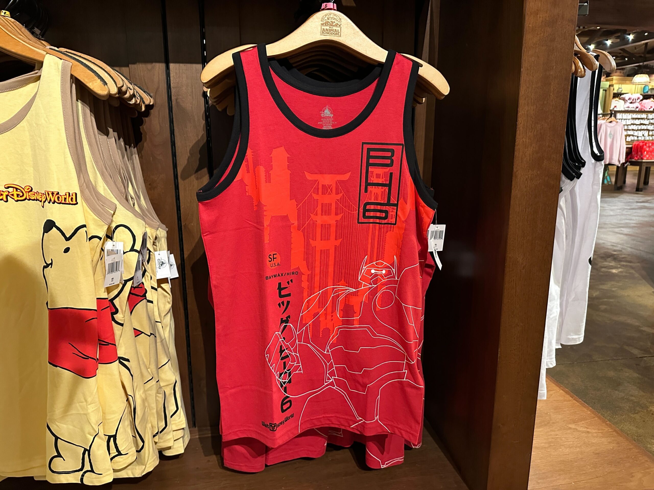 Big Hero 6's Baymax is depicted wearing his armor in white lineart on this red top. In the background is the San Fransokyo skyline, along with symbols for the superhero team.