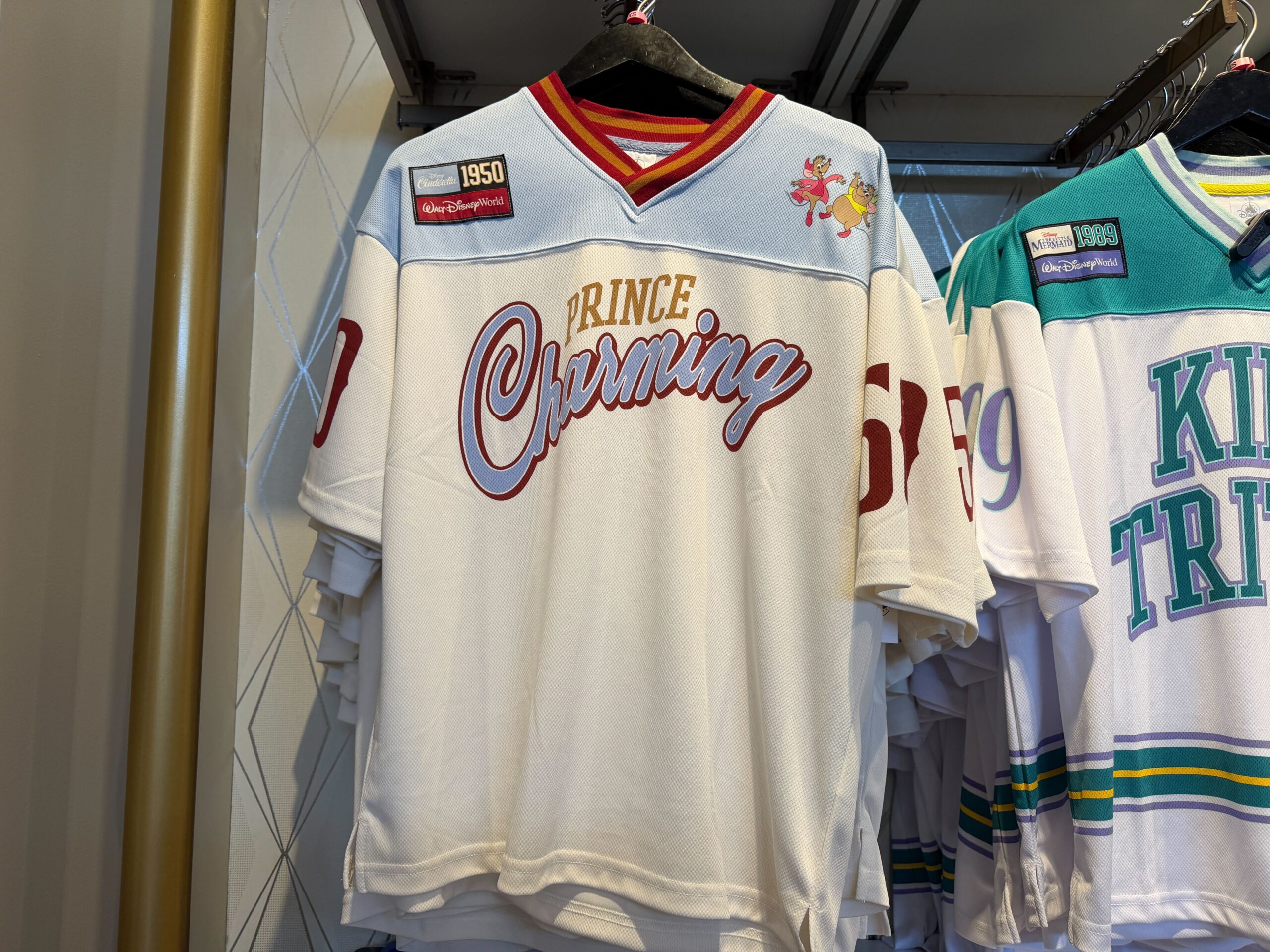 This Cinderella jersey is primarily white with a pale blue shoulder yoke, red-and-gold striped V-neck trim, and red sleeve numbers. The chest reads “Prince Charming” in large flowing script, with “Prince” printed in collegiate lettering above “Charming.” The film's release in 1950 is commemorated with the jersey number, sleeves, and patch the right shoulder.