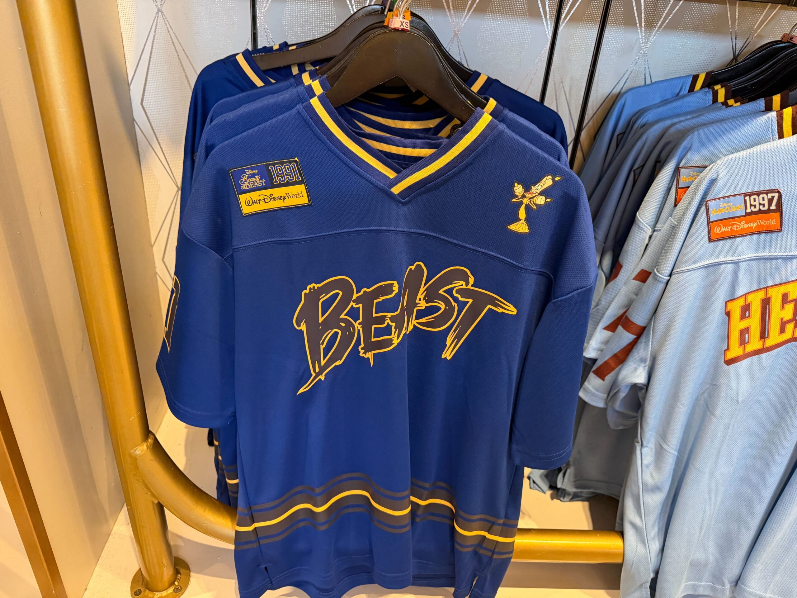 This Beauty and the Beast jersey is a deep royal blue with yellow striping at the collar and hem. The front features “Beast” in a jagged dark script outlined in a gold tone, with a small graphic of Lumiere on the left shoulder. Similar to other items in the collection, the film's release year of 1991 is featured on the back, sleeves, and right shoulder.