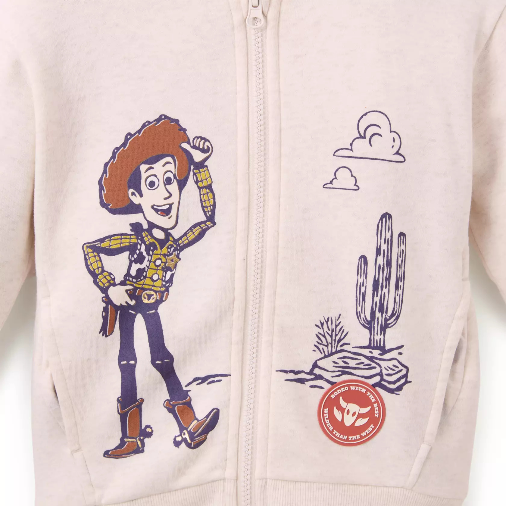 The cream-colored zip hoodie has front screen art of Woody posing in the desert. There is also a rubber patch appliqué with the phrases "Rodeo with the best" and "Wilder than the west." Its pocket lining features screen artwork of Bullseye and Rex.