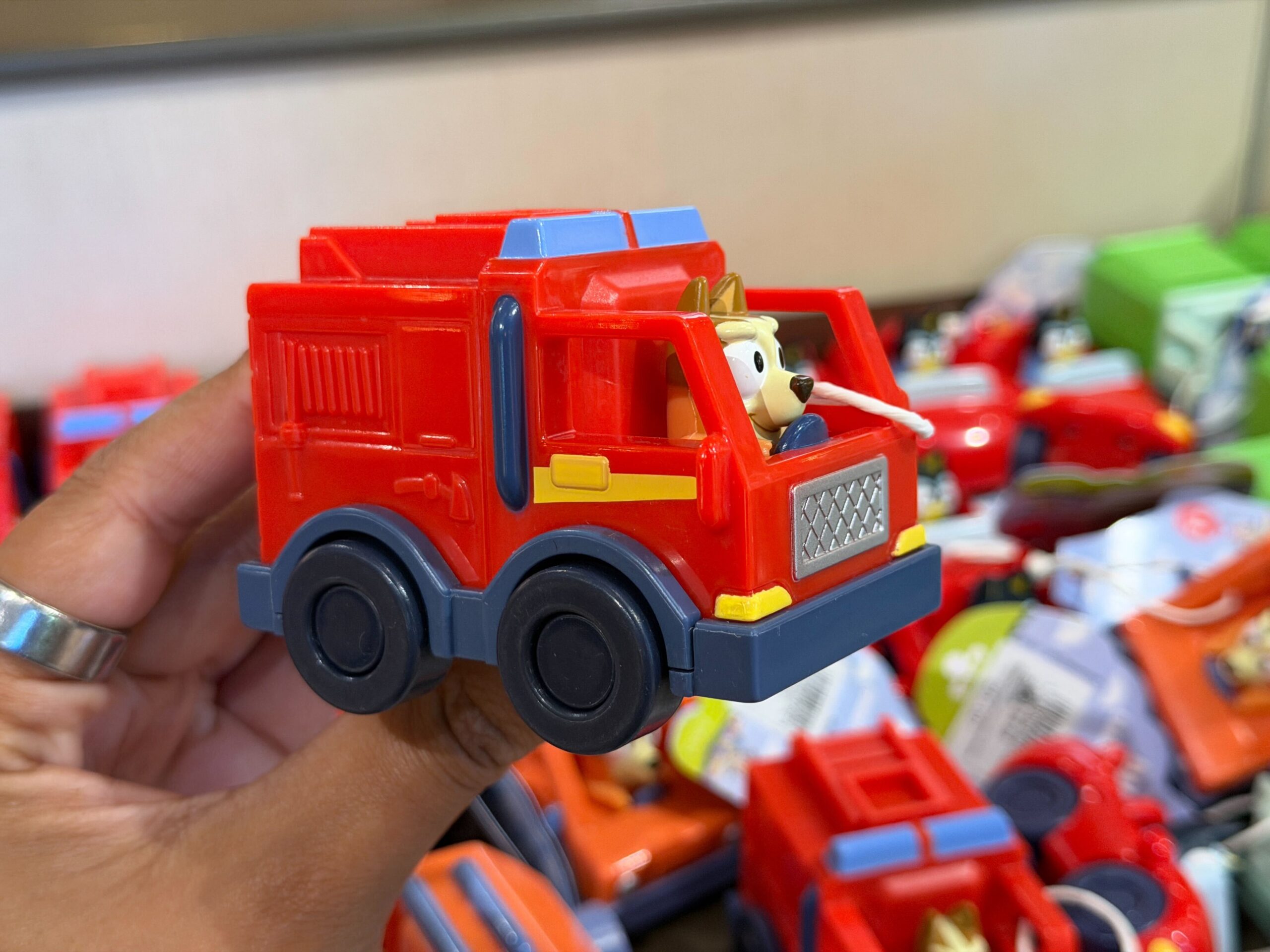 Bluey's younger sister Bingo is depicted driving a red firetruck. These toys have functioning wheels and are recommended for ages 3+.