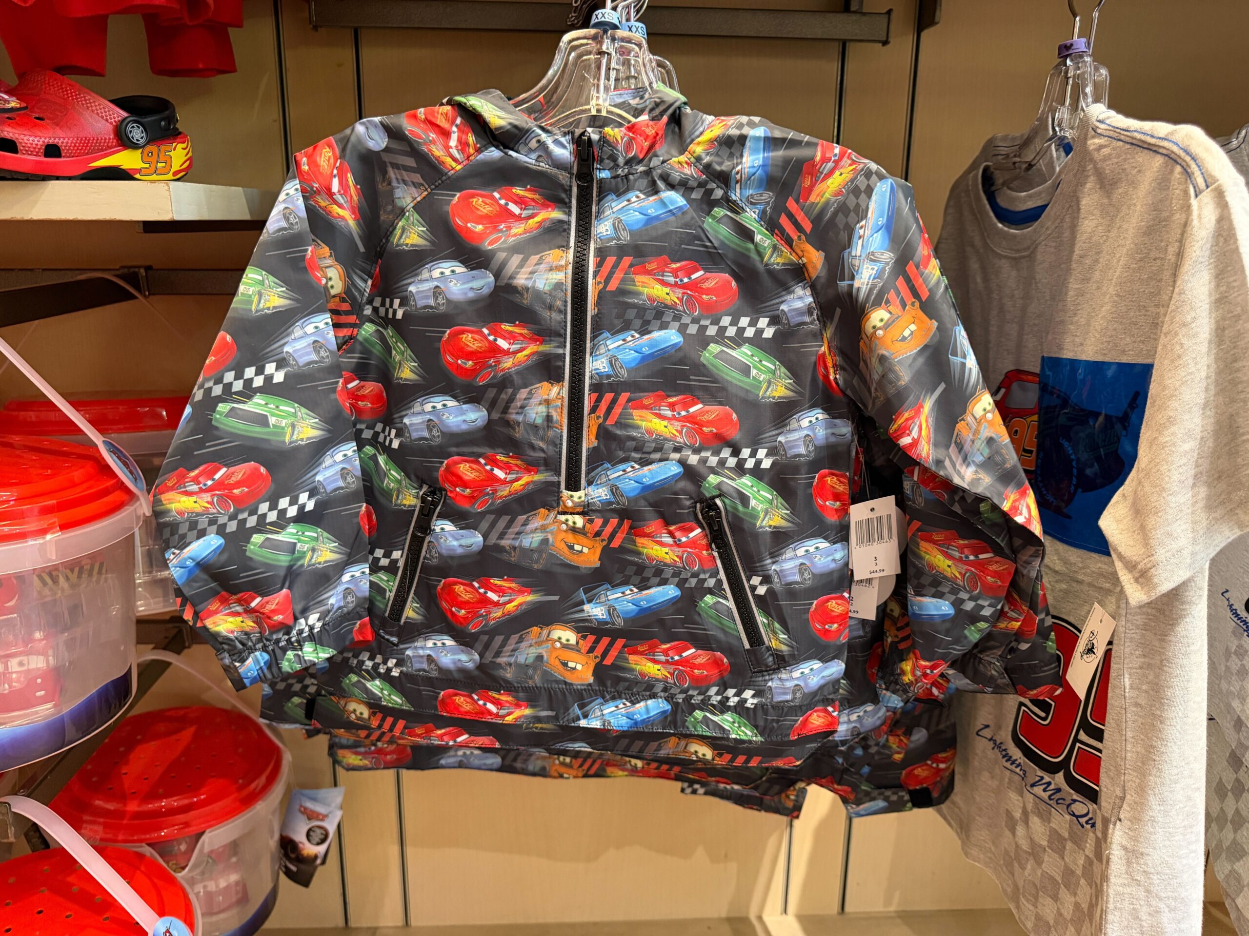 The raincoat is black with an all-over pattern of Lightning, Mater, Sally, Strip Weathers, and Chick Hicks. It has a half-zip front and two zip pockets, plus a hood.