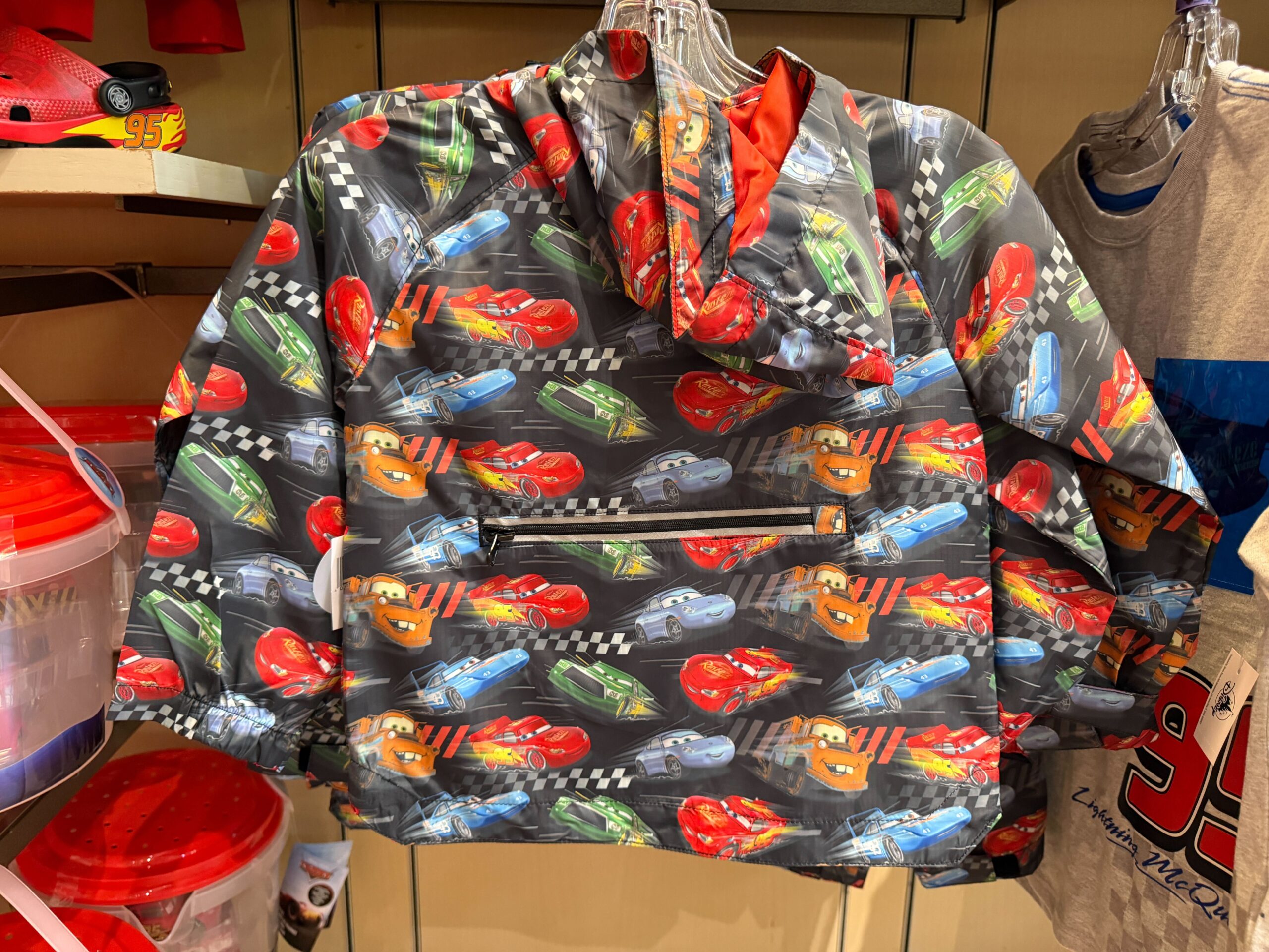 The raincoat is black with an all-over pattern of Lightning, Mater, Sally, Strip Weathers, and Chick Hicks. It has a half-zip front and two zip pockets, plus a hood.