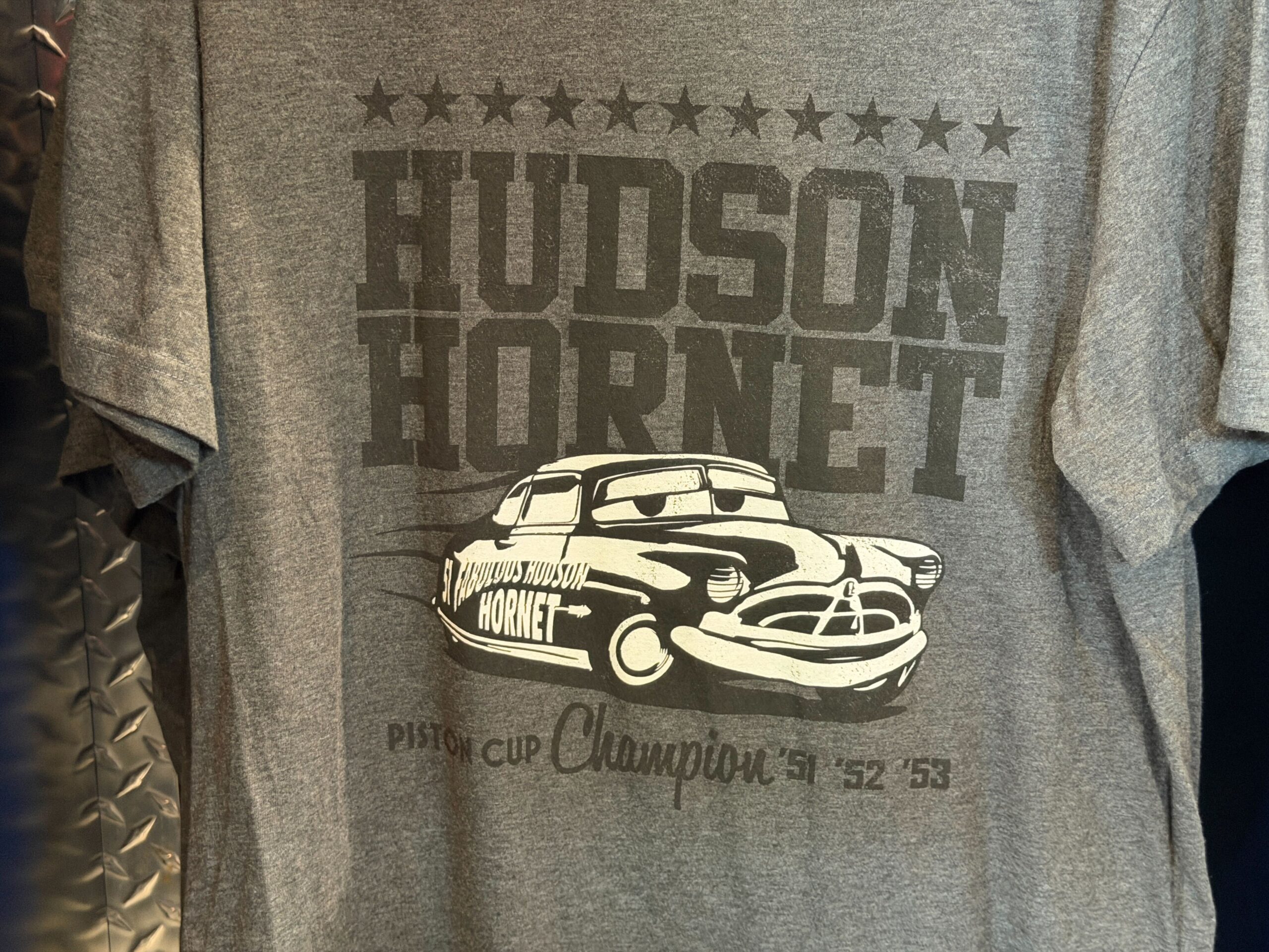 Inspired by the Hudson Hornet a.k.a. Doc Hudson, the famous racecar is pictured in black and white on this heather gray tee. "Hudson Hornet" is in big letters above and behind him, along with a row of stars. Below him is "Piston Cup Champion '51 '52 '53."