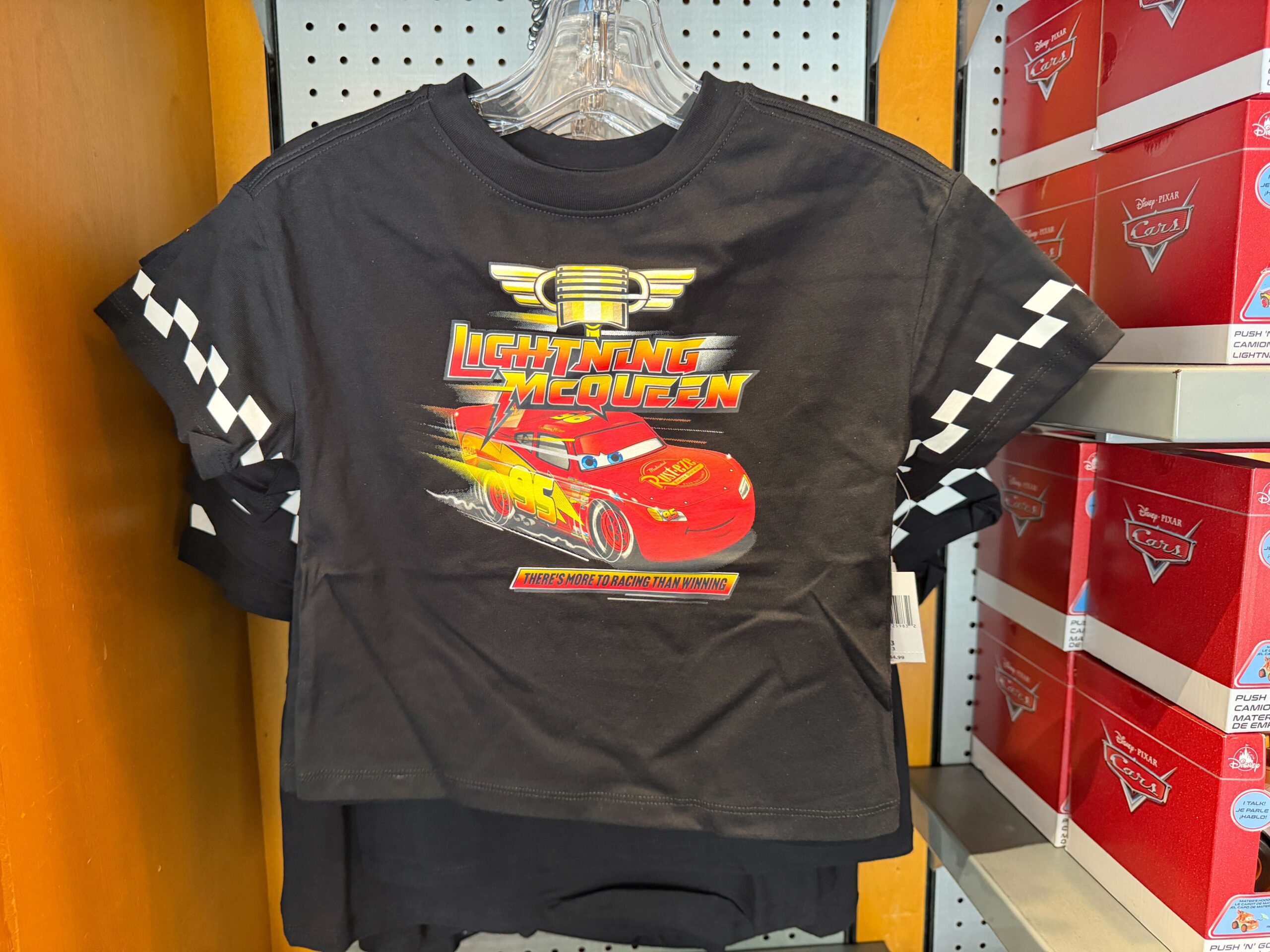 Lightning McQueen is speeding across this shirt. His name and the golden Piston Cup are pictured above him. Below is the phrase, "There's more to racing than winning." The black shirt has white racing stripes on the sleeves.