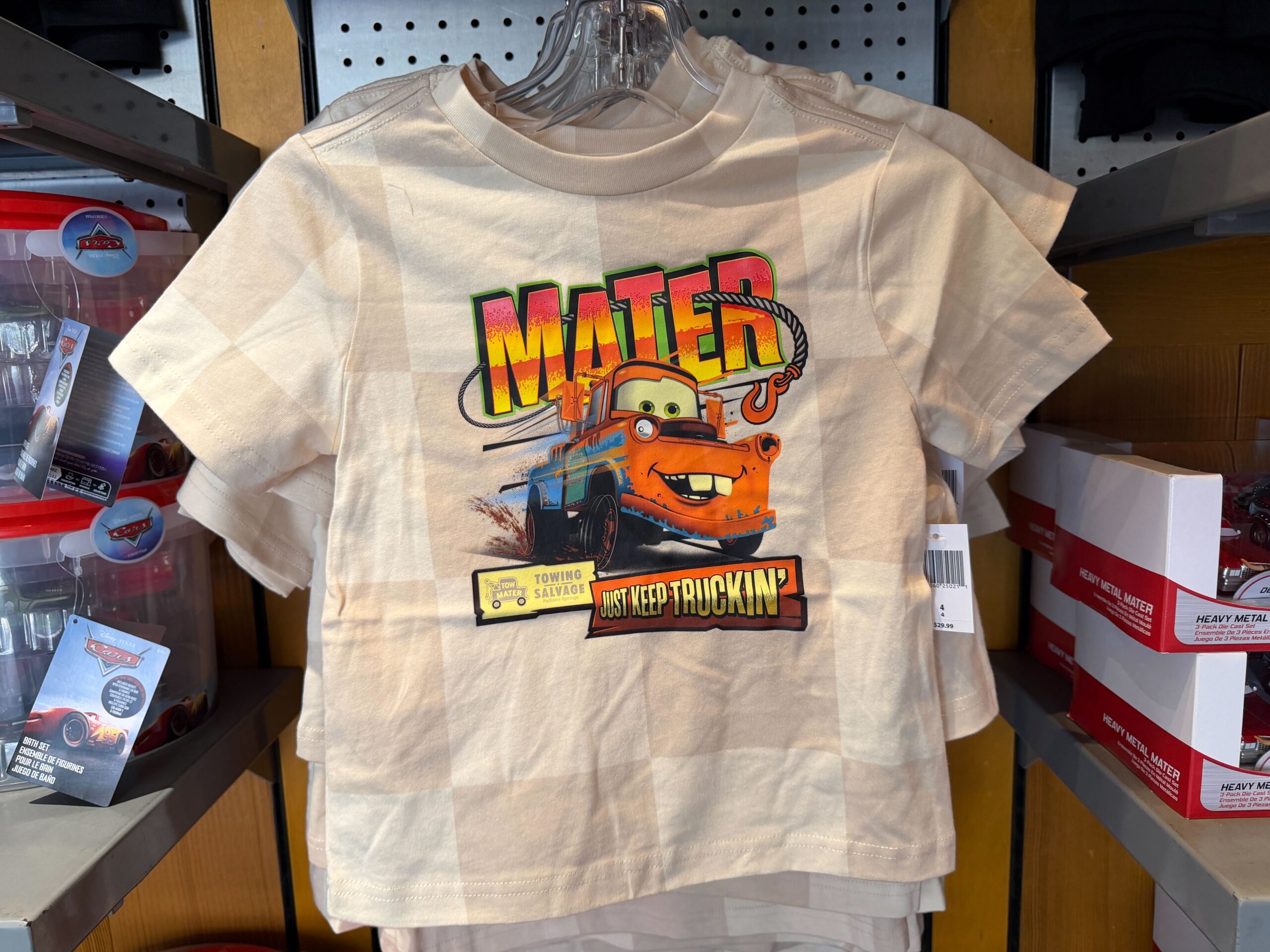 The Mater tee is checkered white and beige. The tow truck is pictured on the front with "Mater" and "Just Keep Truckin'." His tow hook is wrapped around his name and a Towing Salvage sign is in the lower corner.
