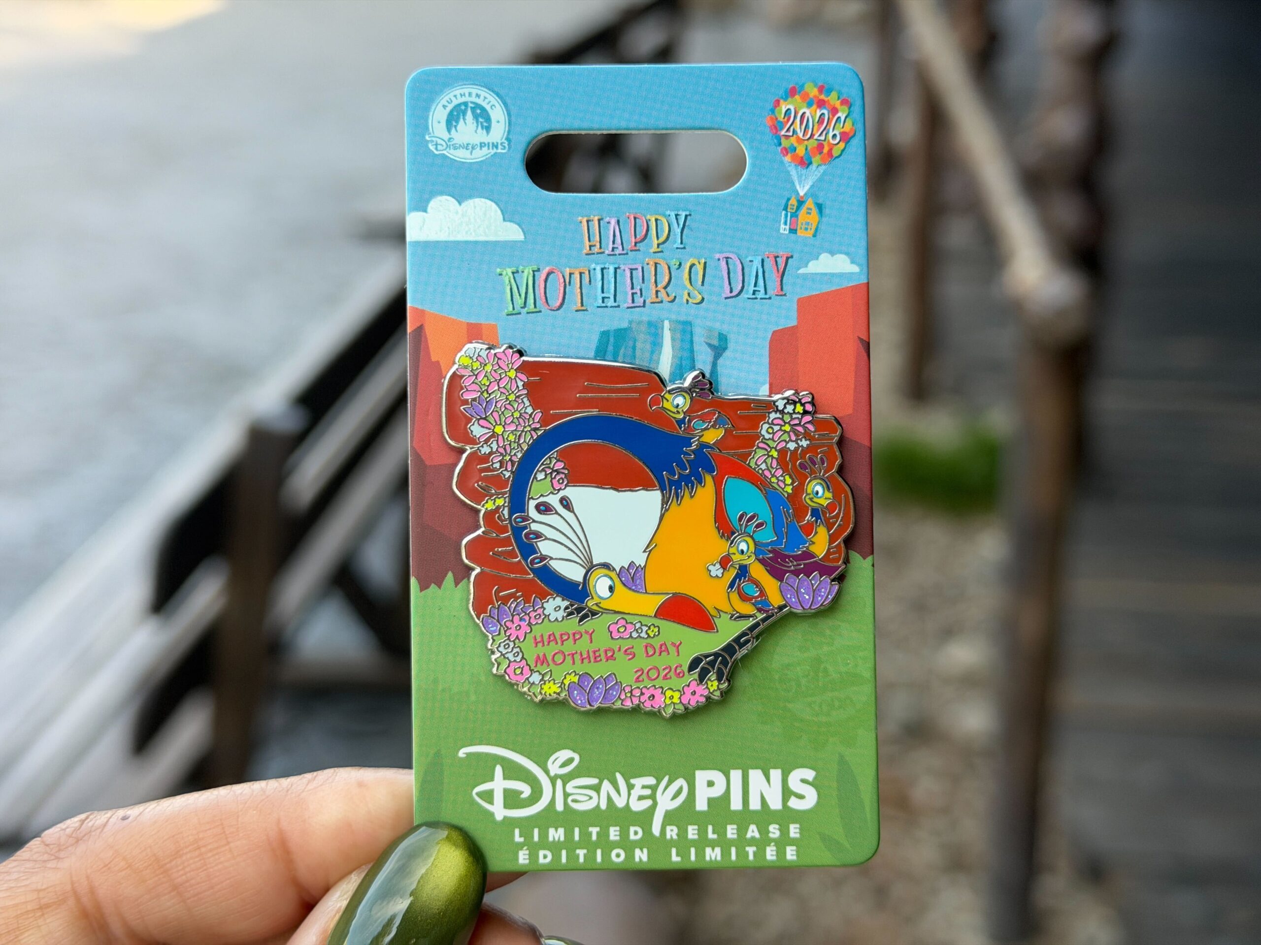 This year's Mother's Day pin features Kevin and her babies from Up. The bird is depicted by a rocky archway with pastel flowers growing around. She has her neck curled down to look at her trio of chicks.