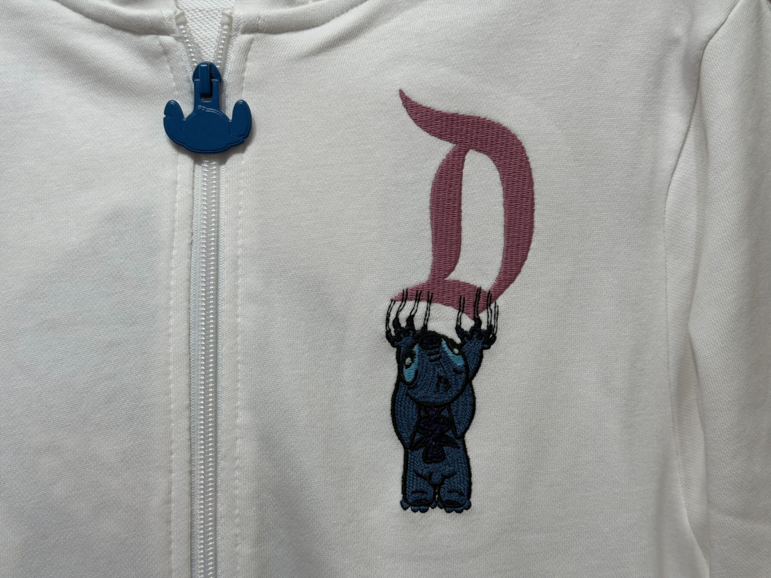 This zip-up hoodie for adults is white and has two front pockets. The front left of the chest depicts Stitch clawing down a pink Disney World “D” logo. The hoodie’s zipper pull is blue and shaped like Stitch’s head.