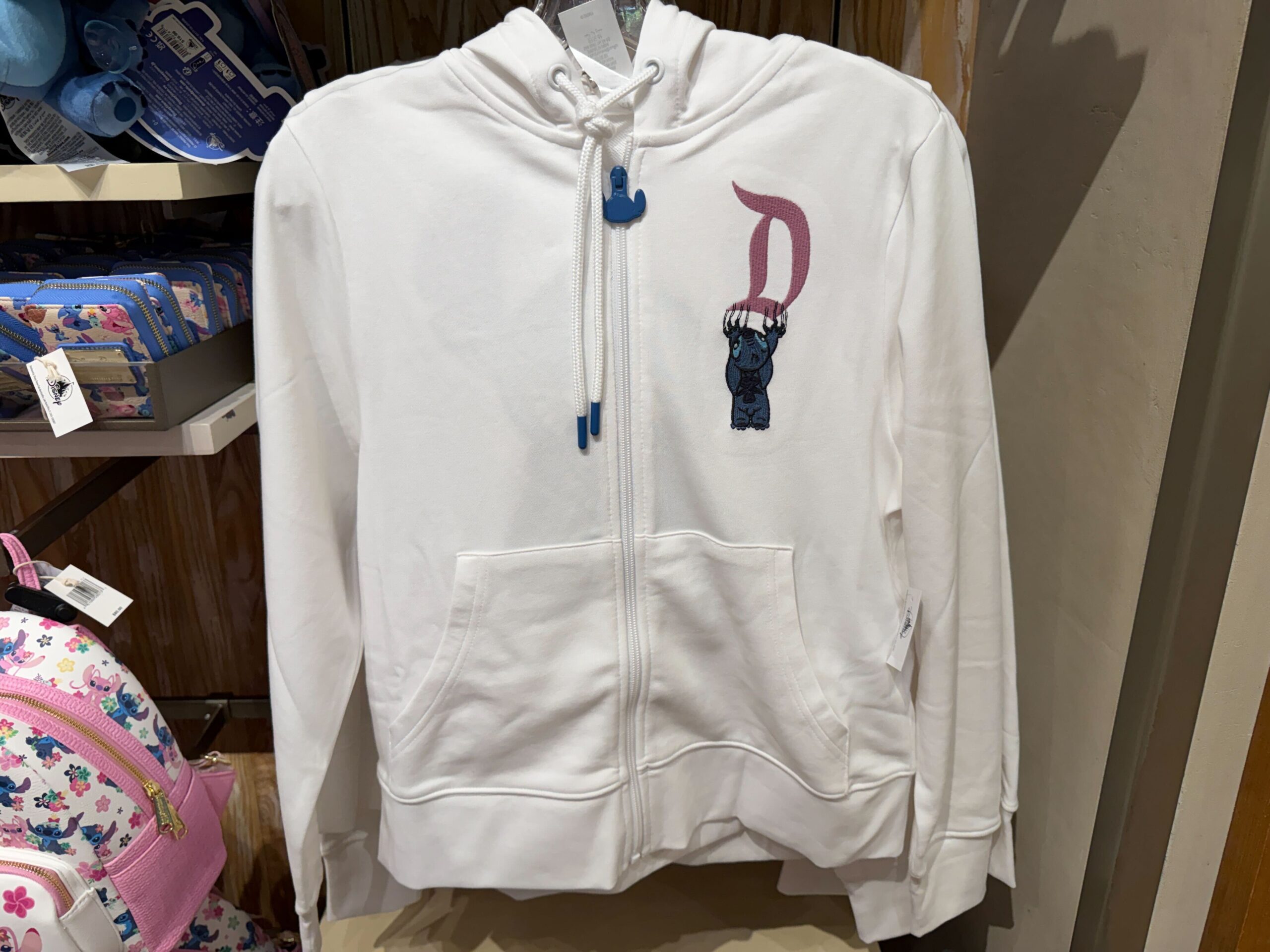 This zip-up hoodie for adults is white and has two front pockets. The front left of the chest depicts Stitch clawing down a pink Disney World “D” logo. The hoodie’s zipper pull is blue and shaped like Stitch’s head.