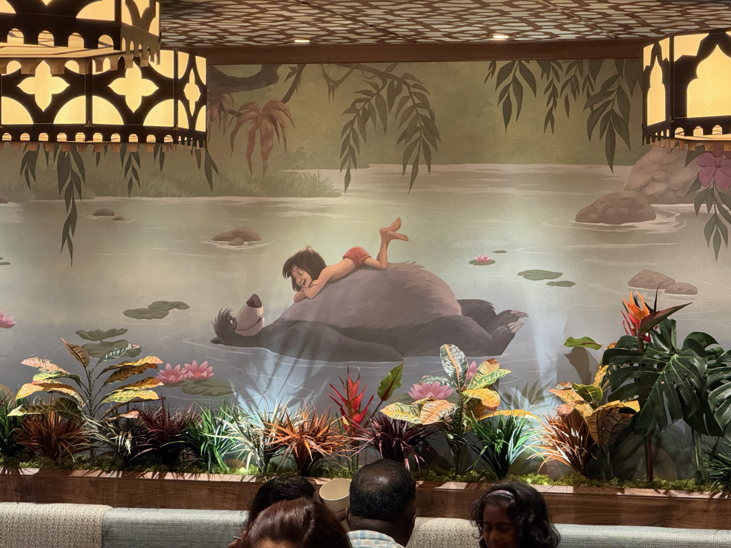 In the back is a large mural scene of Mowgli and Baloo in the water. Dark green panels flank the mural on either side, and a row of flowers are placed in front of the mural.