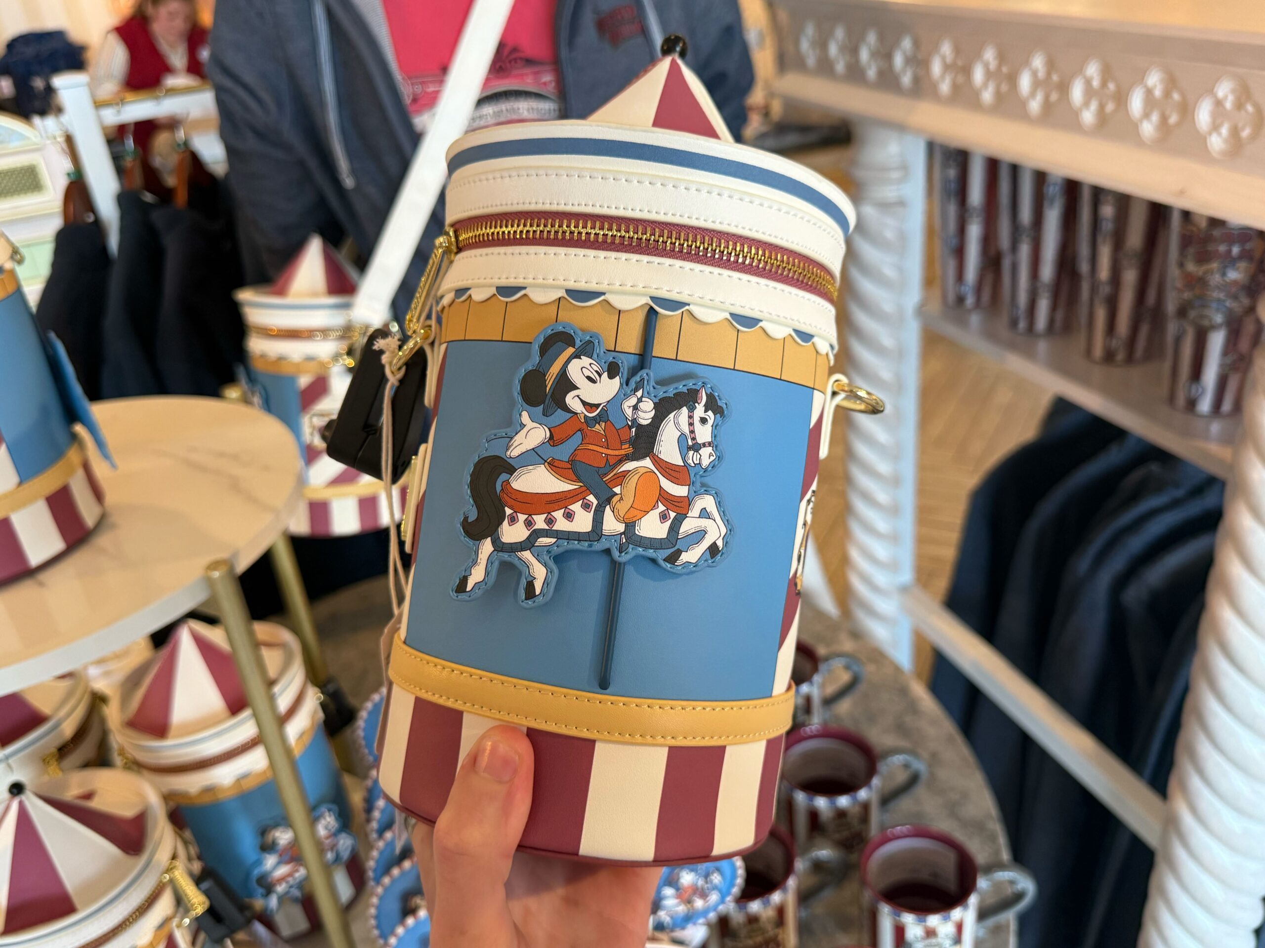 This Loungefly bag is in the shape of a circus tent. It is blue with red and white stripes and yellow details. One one side is an applique of Mickey riding a carousel, and the other side has the BoardWalk wordmark.