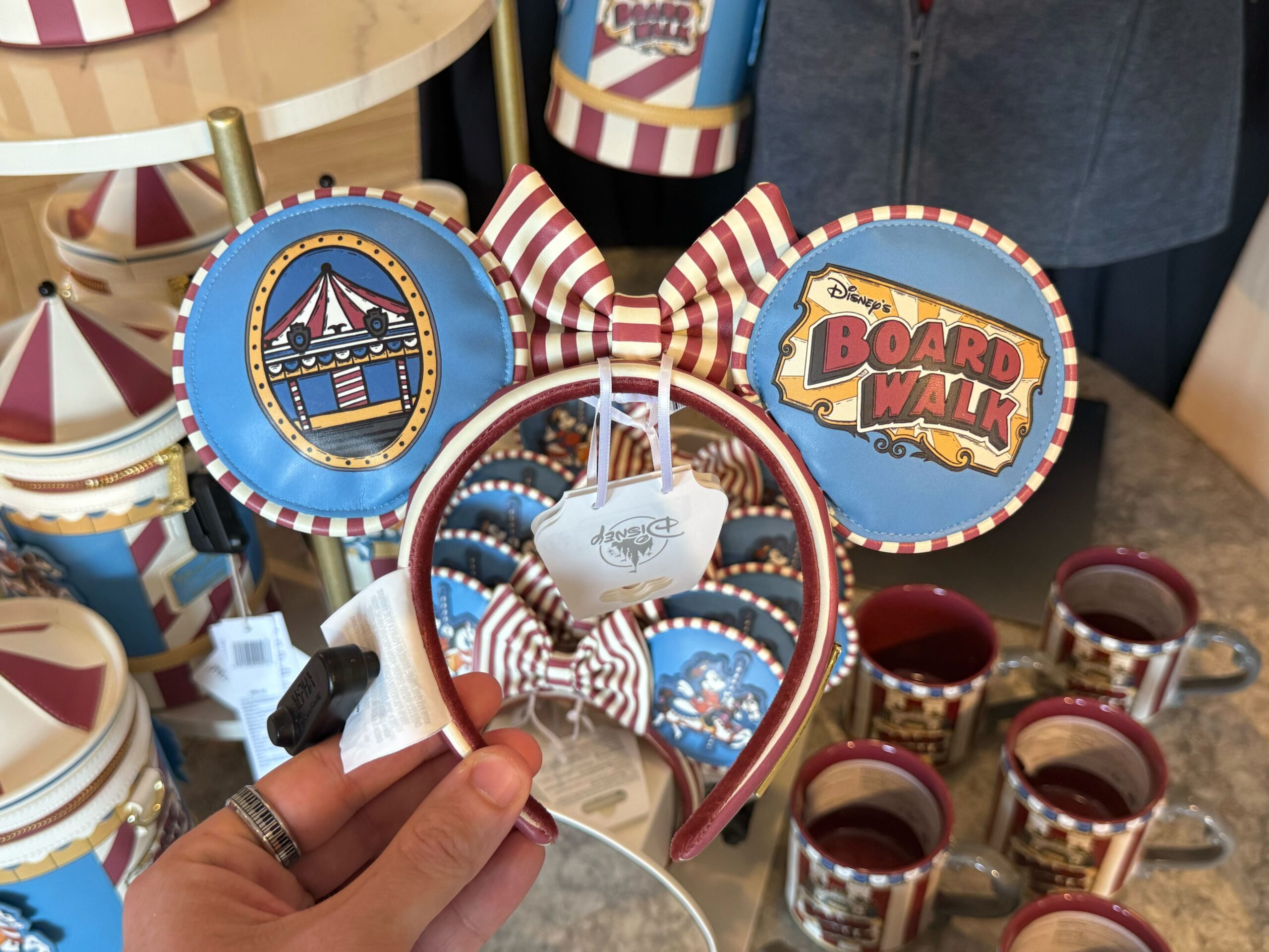 The carousel-themed ear headband features blue ears with Mickey riding the carousel's horse. The back of the ears have an image of the carousel and the resort wordmark. There is a red and white striped bow in the center, and the headband is also striped.