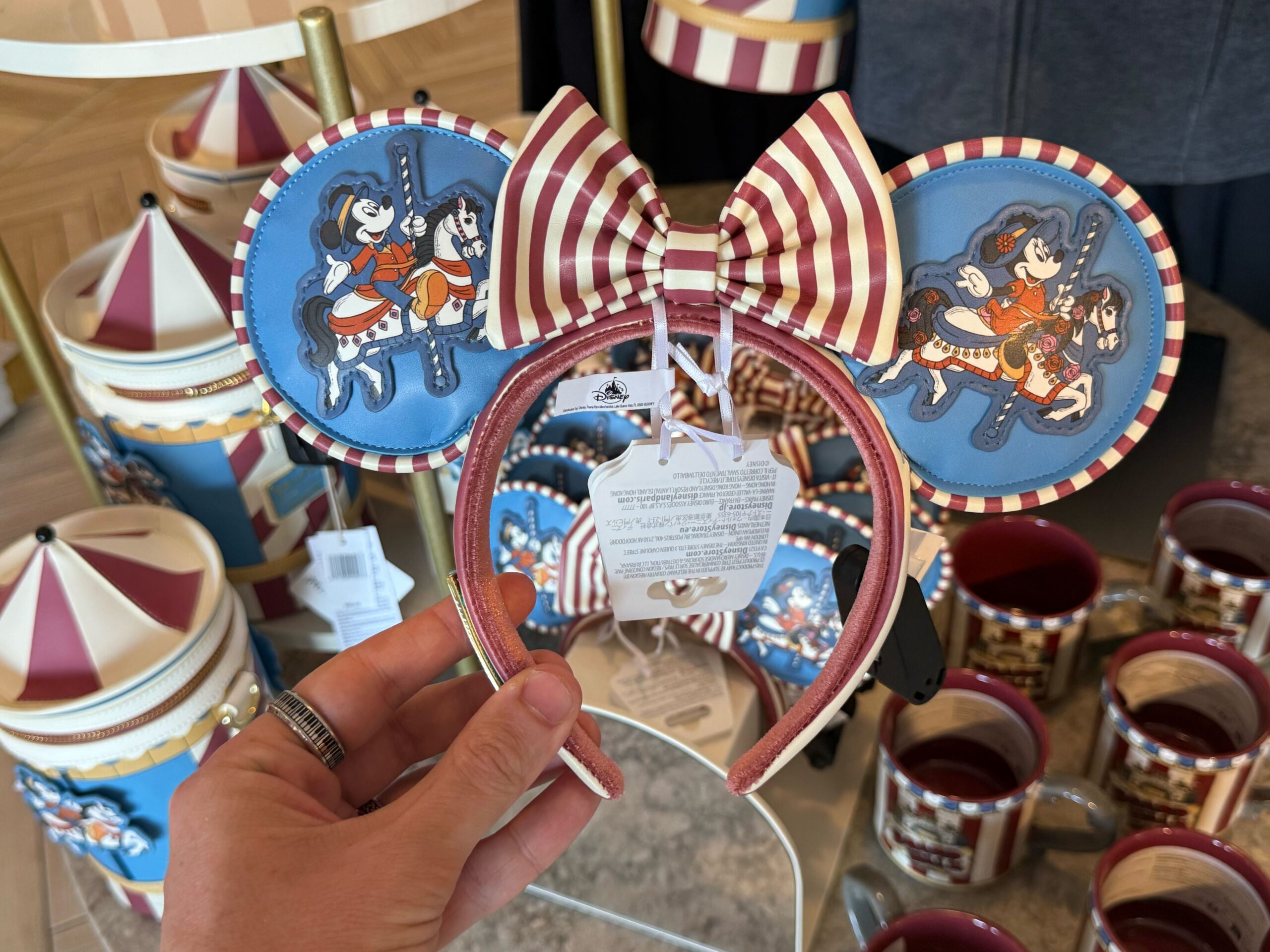 The carousel-themed ear headband features blue ears with Mickey riding the carousel's horse. The back of the ears have an image of the carousel and the resort wordmark. There is a red and white striped bow in the center, and the headband is also striped.