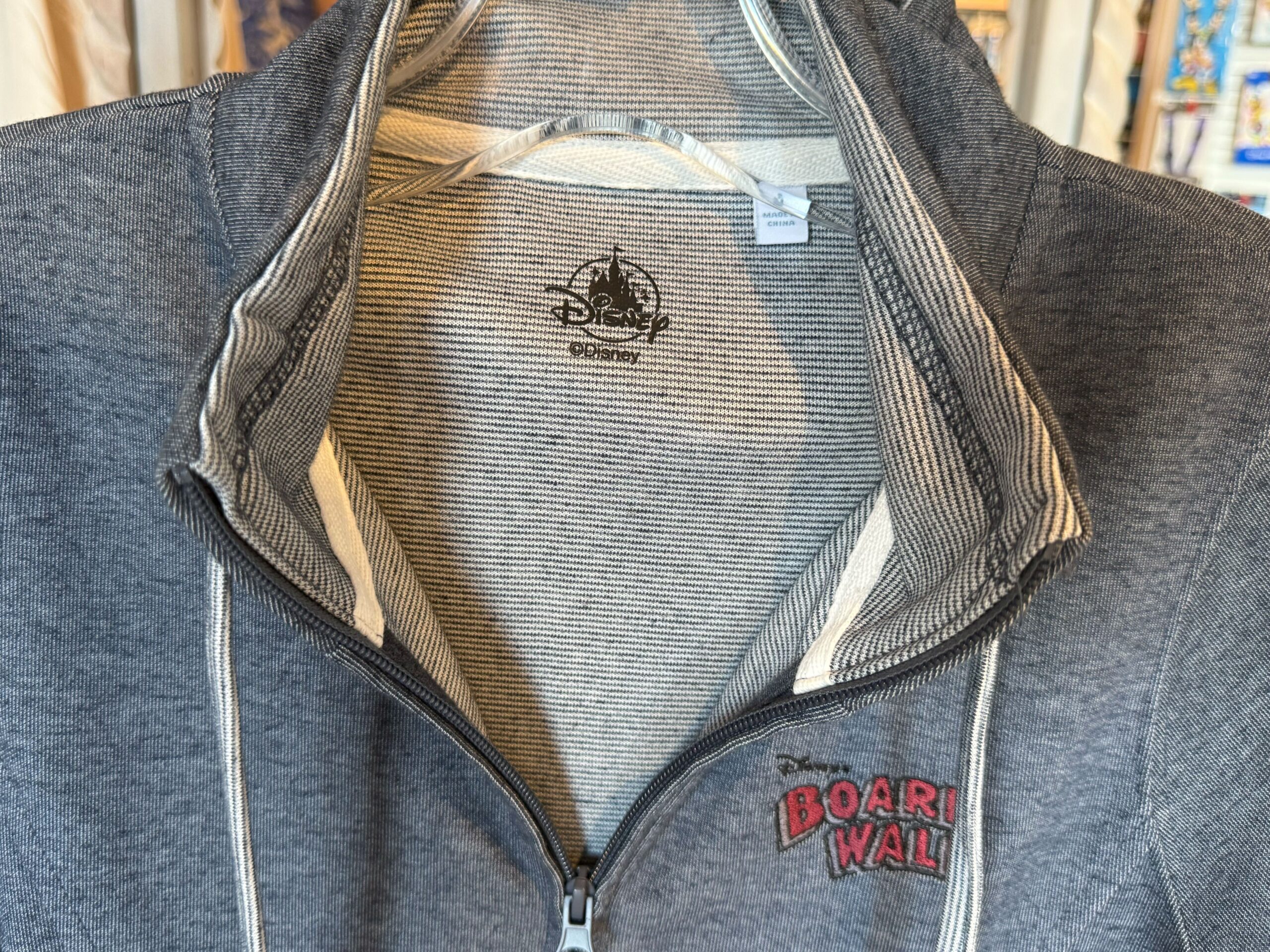 The heather blue zip-up jacket has the resort wordmark on the chest in red text. The strings, sleeve cuffs, and interior lining are a light blue striped color.