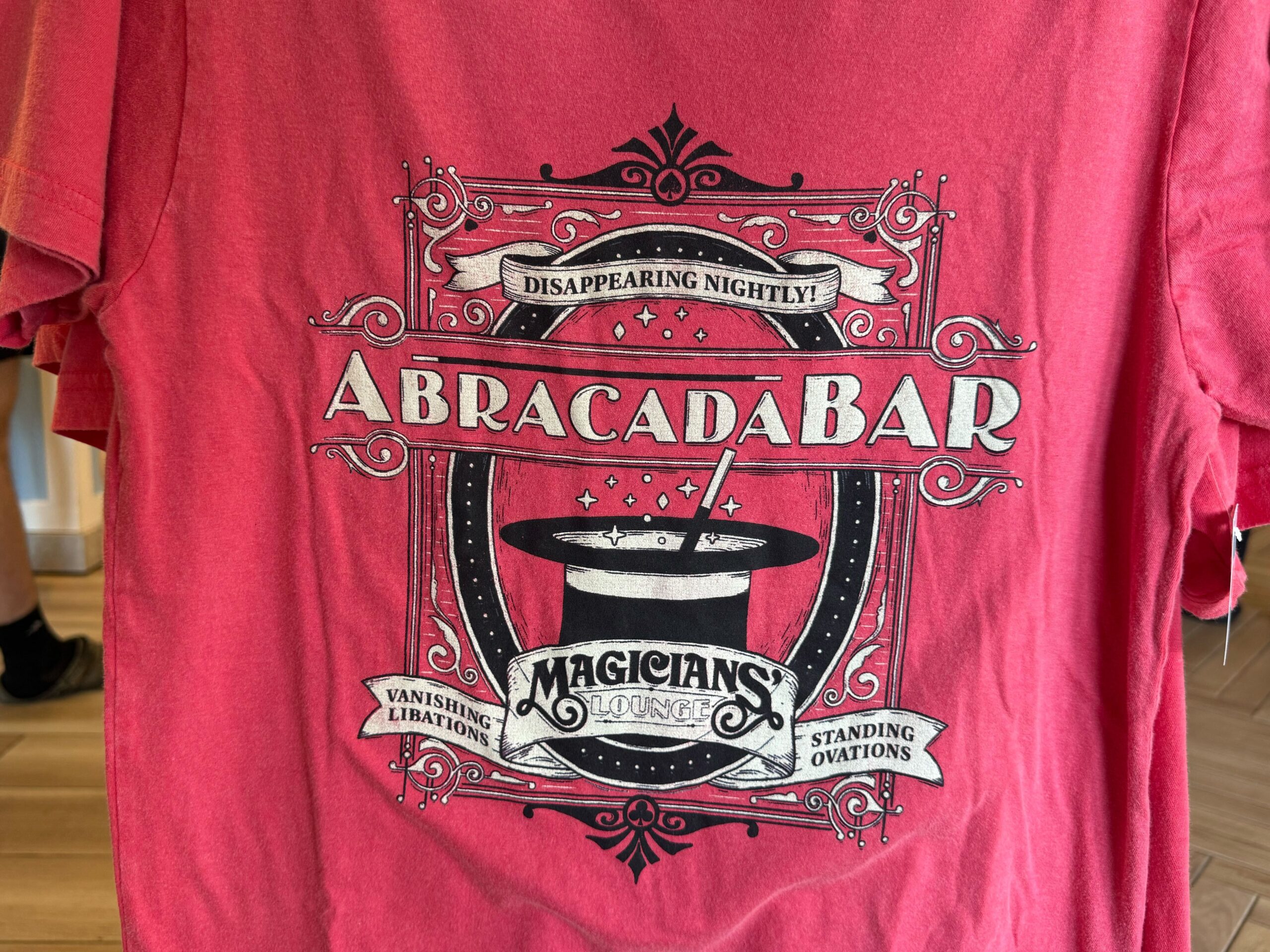 The resort bar, called Abracadabar, is featured on this red T-shirt. The text reads "Disappearing Nightly" on a banner at the top and "Magician's Lounge" on the bottom banner. The sides read "Vanishing Libations" and "Standing Ovations." A magician's hat is in the center of the image.