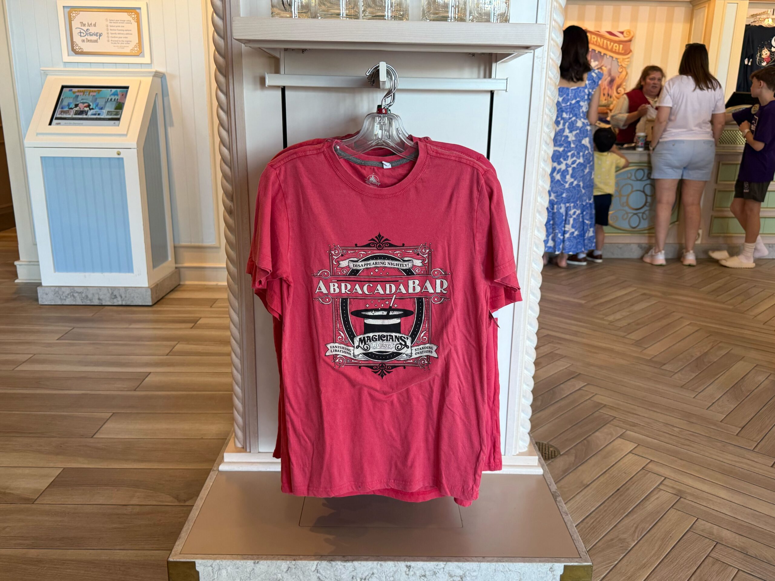 The resort bar, called Abracadabar, is featured on this red T-shirt. The text reads "Disappearing Nightly" on a banner at the top and "Magician's Lounge" on the bottom banner. The sides read "Vanishing Libations" and "Standing Ovations." A magician's hat is in the center of the image.