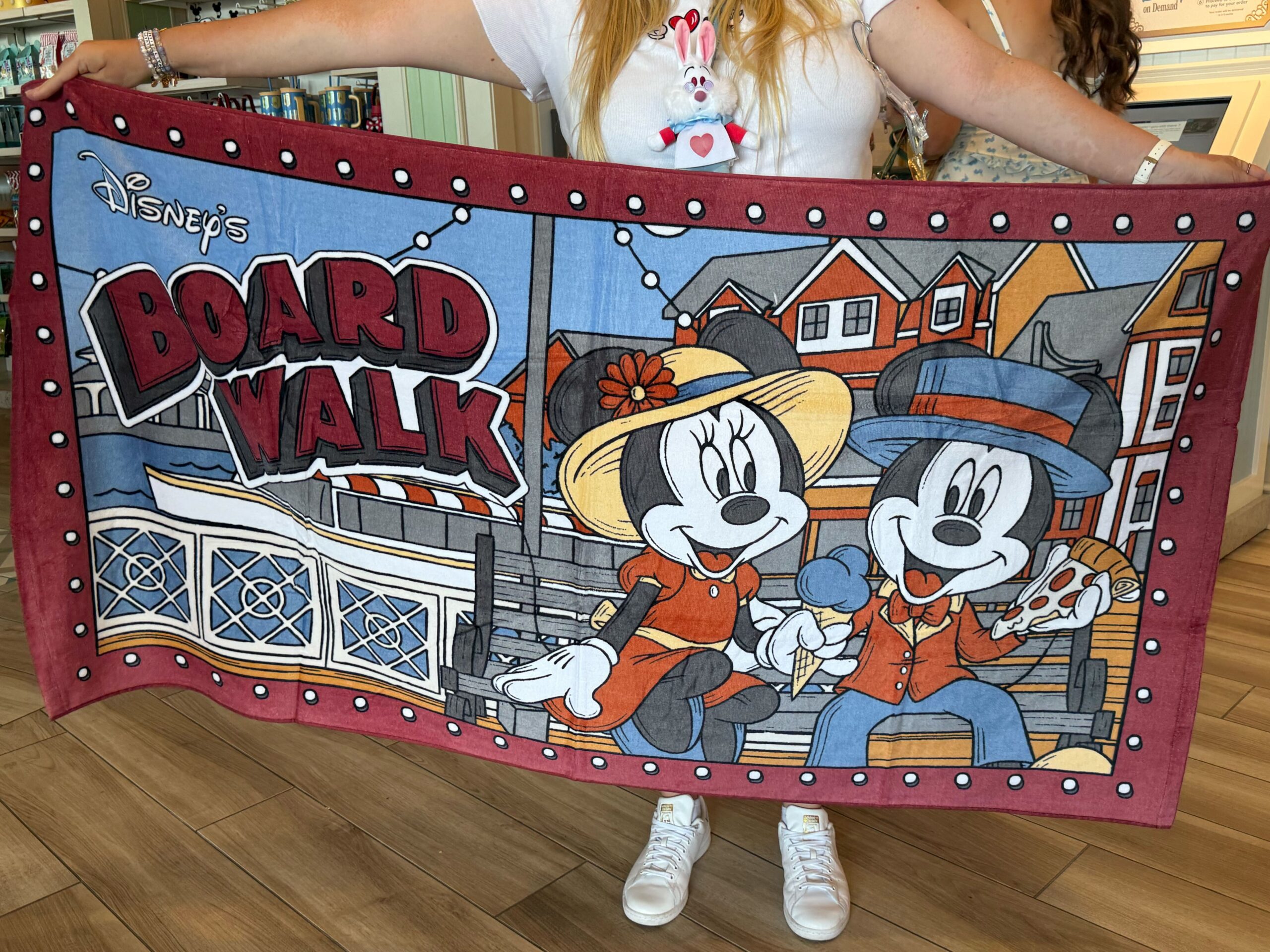 This large beach towel has an image of Mickey and Minnie sitting on a bench enjoying Boardwalk treats. Crescent Lake and resort buildings are in the back, and the BoardWalk wordmark is in the upper left corner. The red border has white lights.