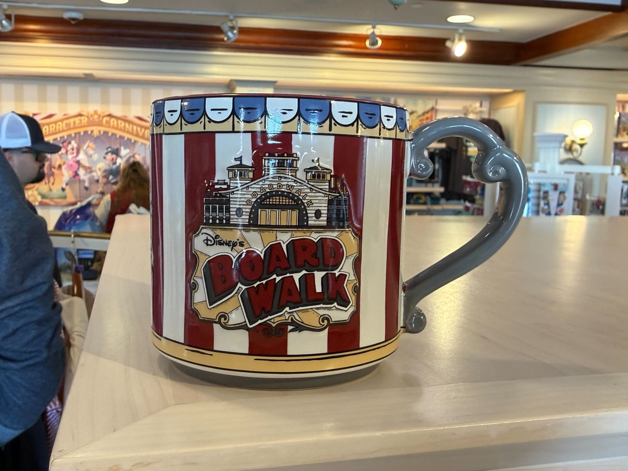 The mug is similarly designed to other items in this collection—with red and white stripes and an image of Mickey riding on the carousel's horse. Another image features the front of the resort and the wordmark. A blue and white banner lines the top. The interior is the same dark red color, and a Mickey icon is stamped on the bottom. The mug has a vintage style to match the feel of the resort and collection.