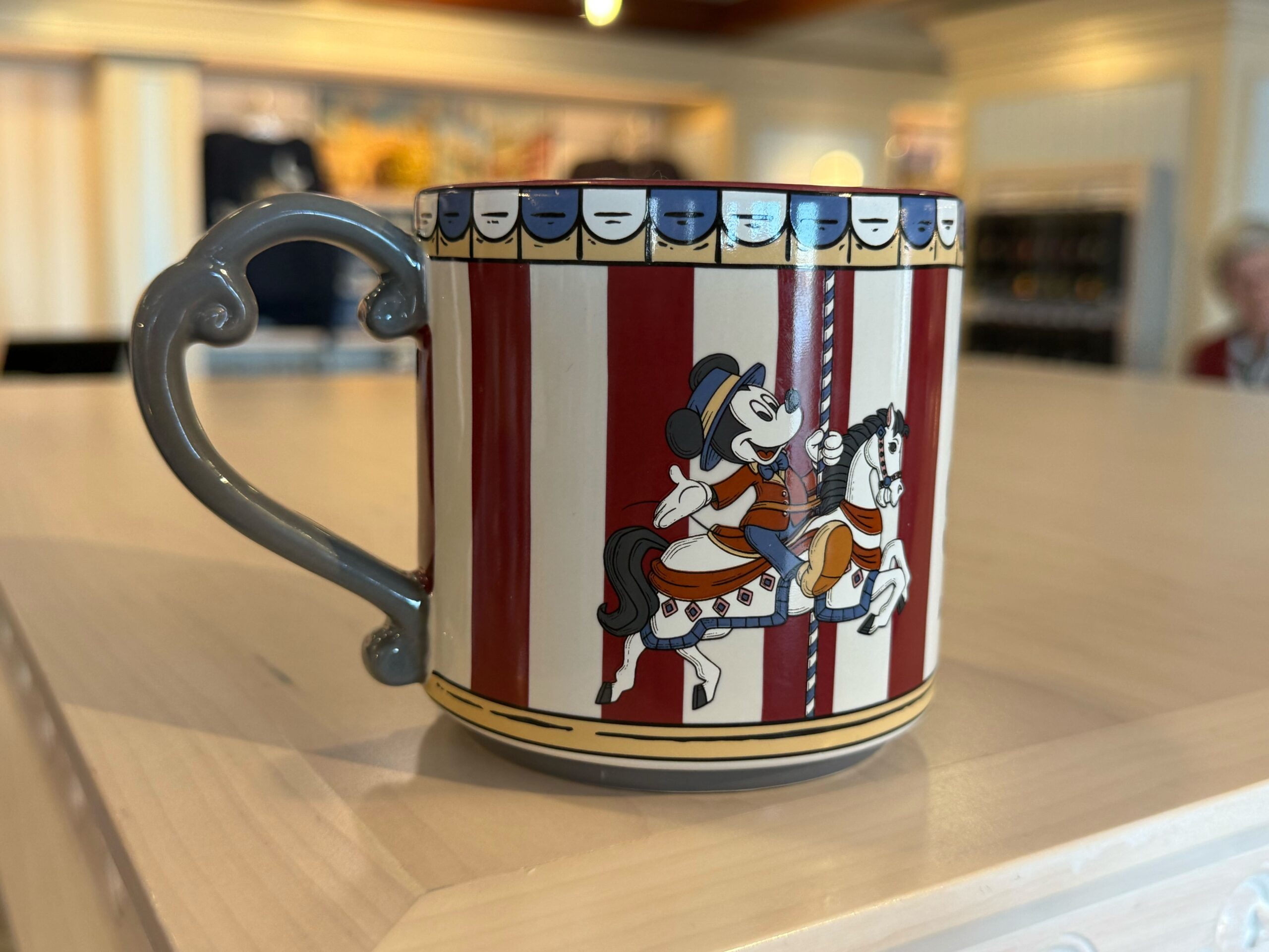 The mug is similarly designed to other items in this collection—with red and white stripes and an image of Mickey riding on the carousel's horse. Another image features the front of the resort and the wordmark. A blue and white banner lines the top. The interior is the same dark red color, and a Mickey icon is stamped on the bottom. The mug has a vintage style to match the feel of the resort and collection.
