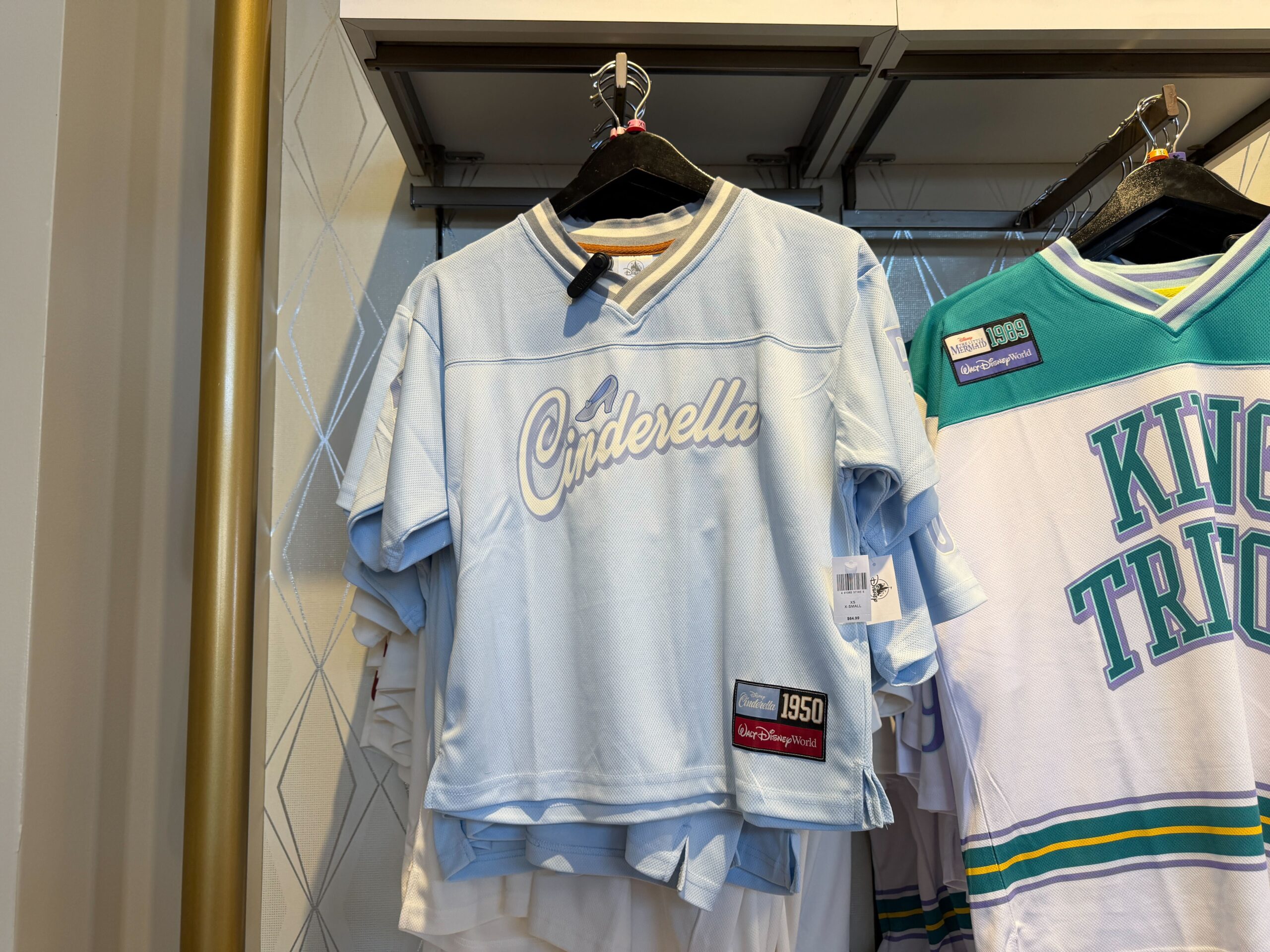 This Cinderella jersey is primarily a light blue color with gray and white striped V-neck trim, and a slightly darker blue on the sleeve number. The chest reads “Cinderella” in large flowing script, with her shoe above the text.