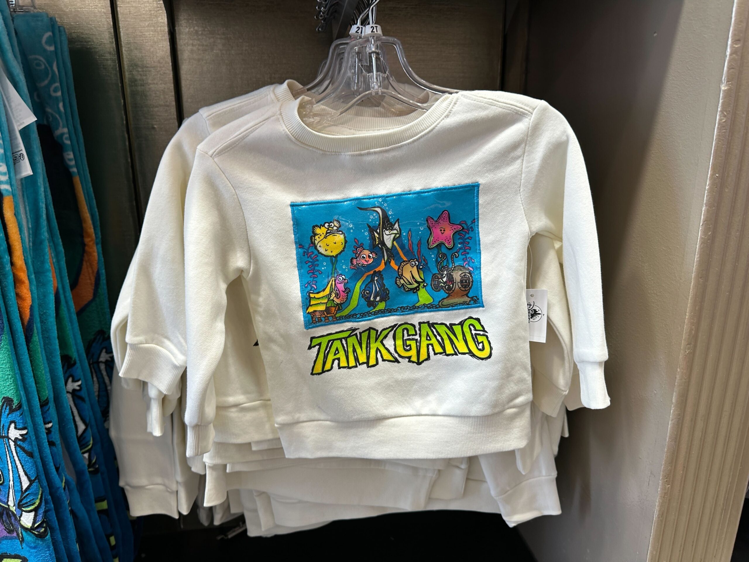 This youth sweater is white with an image of Mount Wannahockaloogie, the toy volcano from the dentist’s tank. Around the volcano are the various fish: Nemo, Gill, Bloat, Peach, Bubbles, Gurgle, Deb, and Jacques. "Tank Gang" text is at the bottom. There is a beach towel with the same design.