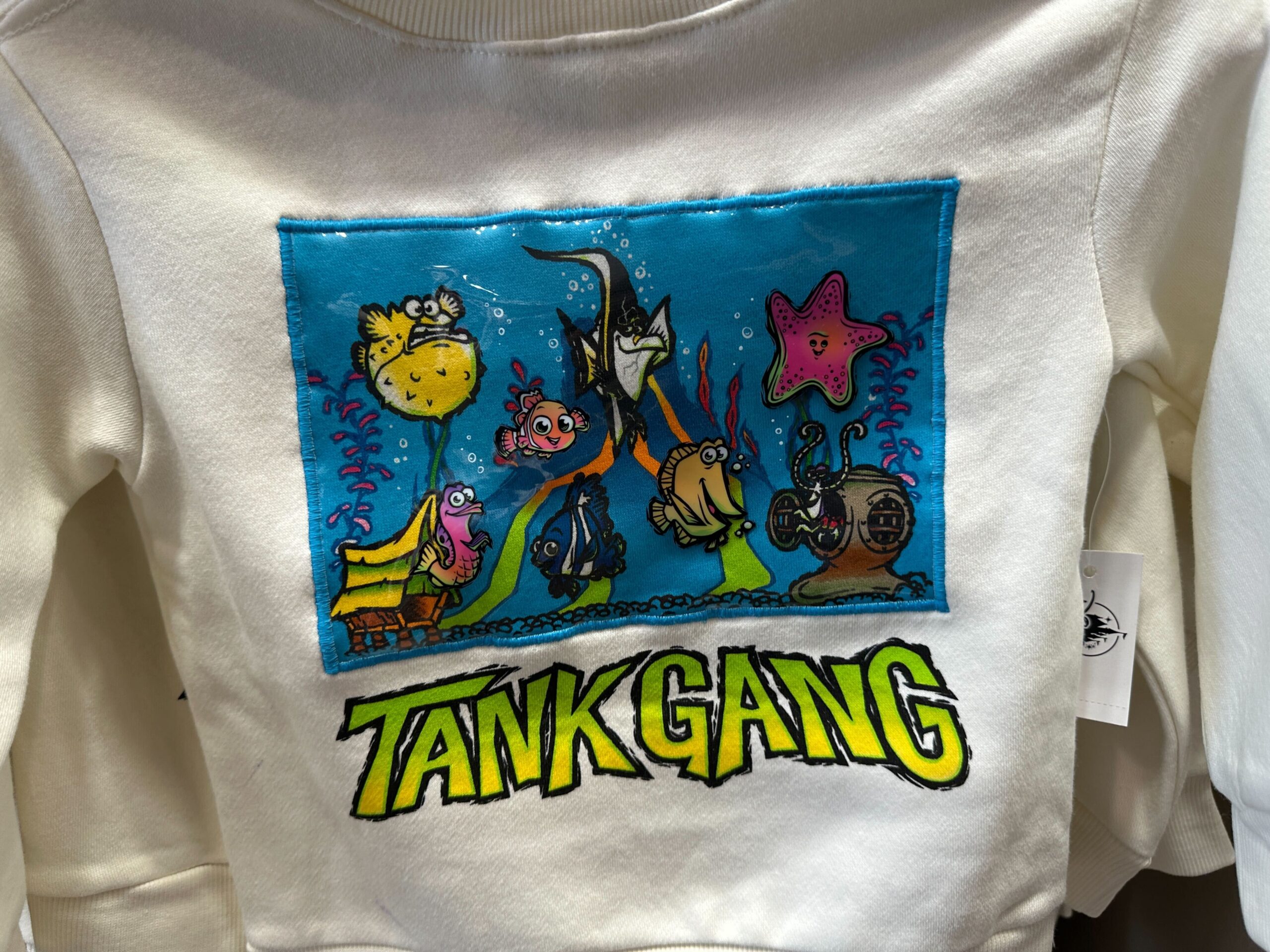 This youth sweater is white with an image of Mount Wannahockaloogie, the toy volcano from the dentist’s tank. Around the volcano are the various fish: Nemo, Gill, Bloat, Peach, Bubbles, Gurgle, Deb, and Jacques. "Tank Gang" text is at the bottom. There is a beach towel with the same design.