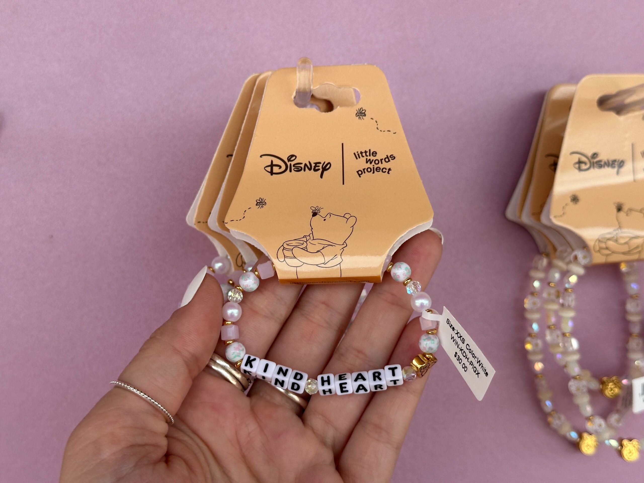 The block letters read “Kind Heart” with a gold Piglet bead on one end. At the back of the bracelet is a dangling “Hunny” pot charm. The beads in this bracelet are white with floral accents, pink, gold, ivory, or pearlescent.