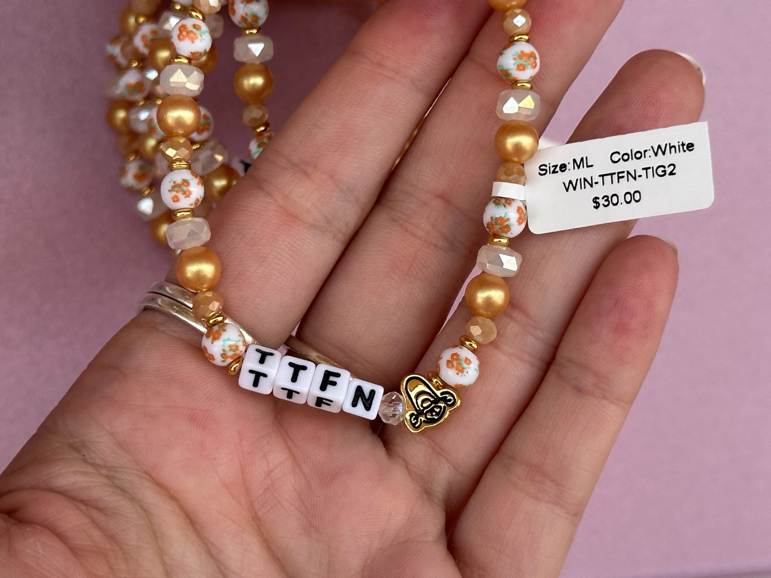 The block letters read “TTFN” (ta ta for now) with a gold Tigger bead on one end. At the back of the bracelet is a dangling “Hunny” pot charm. These beads are in shades of orange, gold, and white with orange floral accents.