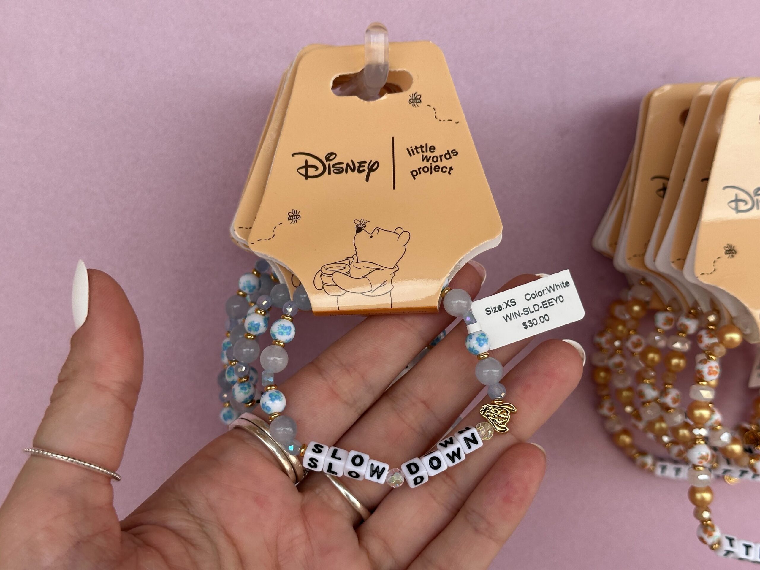The block letters read “Slow Down” with a gold Eeyore bead on one end. At the back of the bracelet is a dangling “Hunny” pot charm. These beads are in shades of blue, gold, and white with blue floral accents.