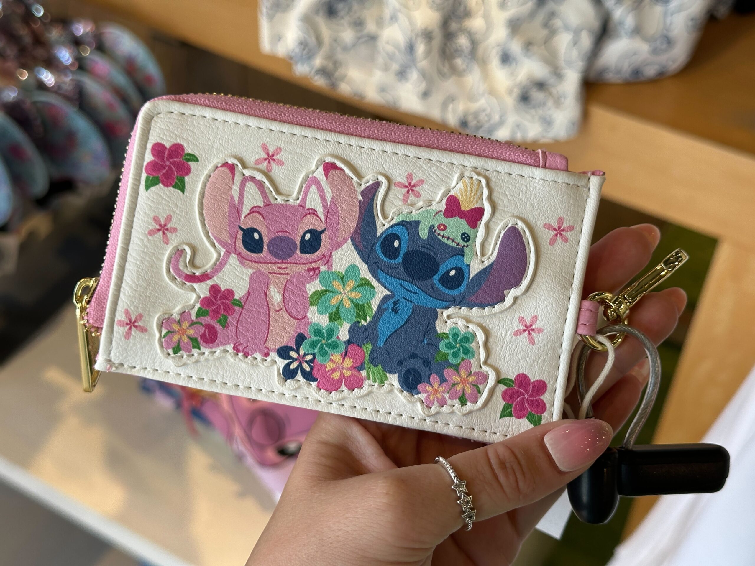 This white Loungefly wallet has a design of Angel and Stitch sitting side-by-side while multicolored tropical flowers surround them. Scrump sits on Stitch's head.