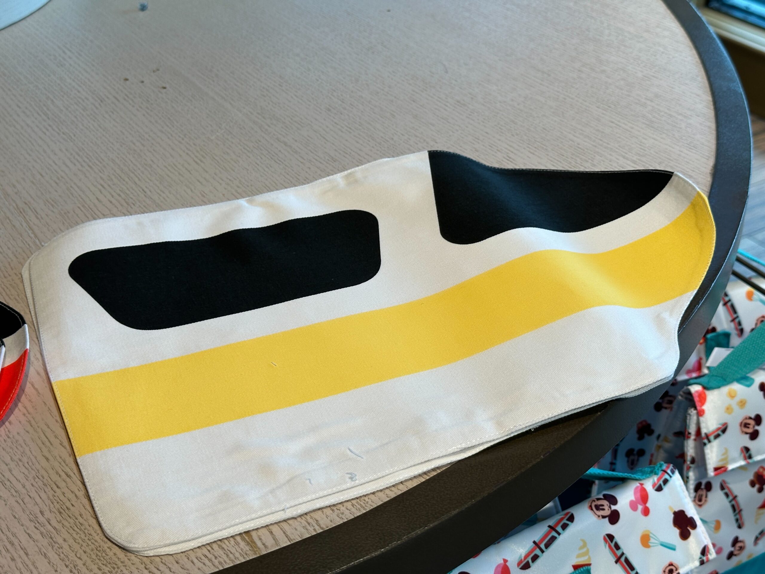 The fabric placemat is in the shape of a Monorail car, with the blacked out windows and yellow stripe that runs along the side.