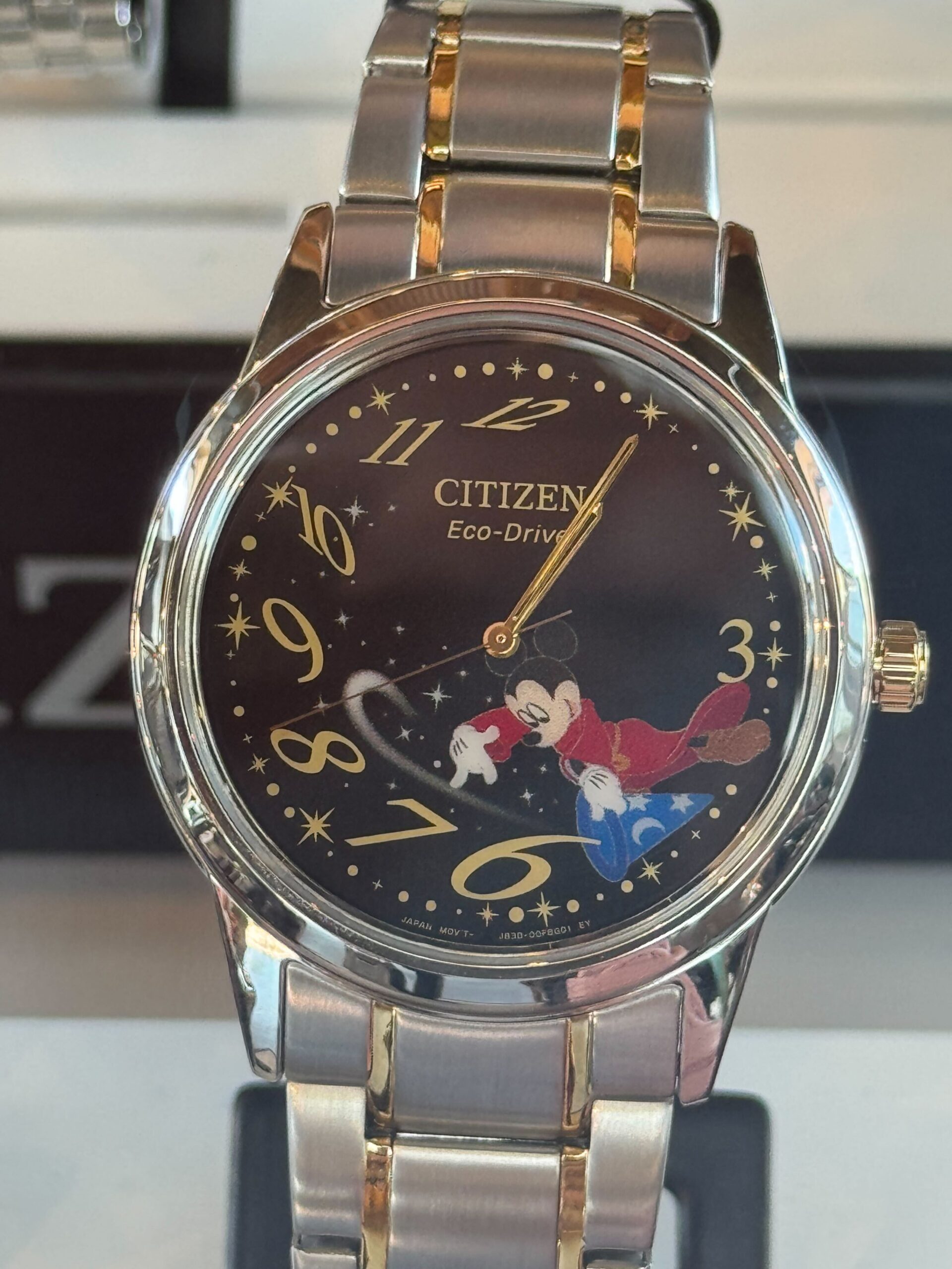 The Sorcerer Mickey watch by Citizen features a silver-tone round case paired with a brushed strap. The design of the black dial shows Sorcerer Mickey positioned between the 3 and 6 numerals and pointing toward the 6. There are gold-tone details on the strap and crown, as well as the numbers on the face.