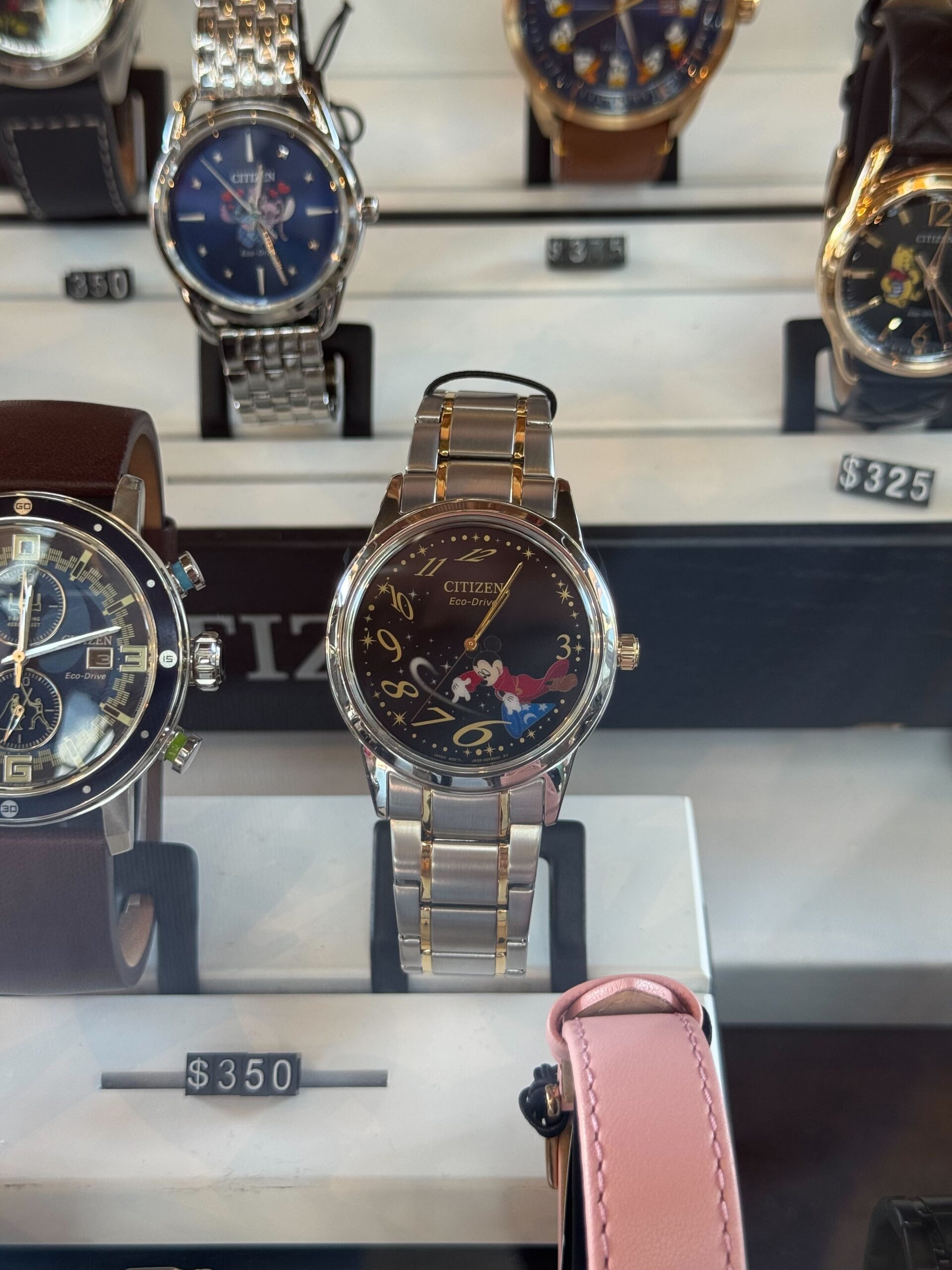 The Sorcerer Mickey watch by Citizen features a silver-tone round case paired with a brushed strap. The design of the black dial shows Sorcerer Mickey positioned between the 3 and 6 numerals and pointing toward the 6. There are gold-tone details on the strap and crown, as well as the numbers on the face.