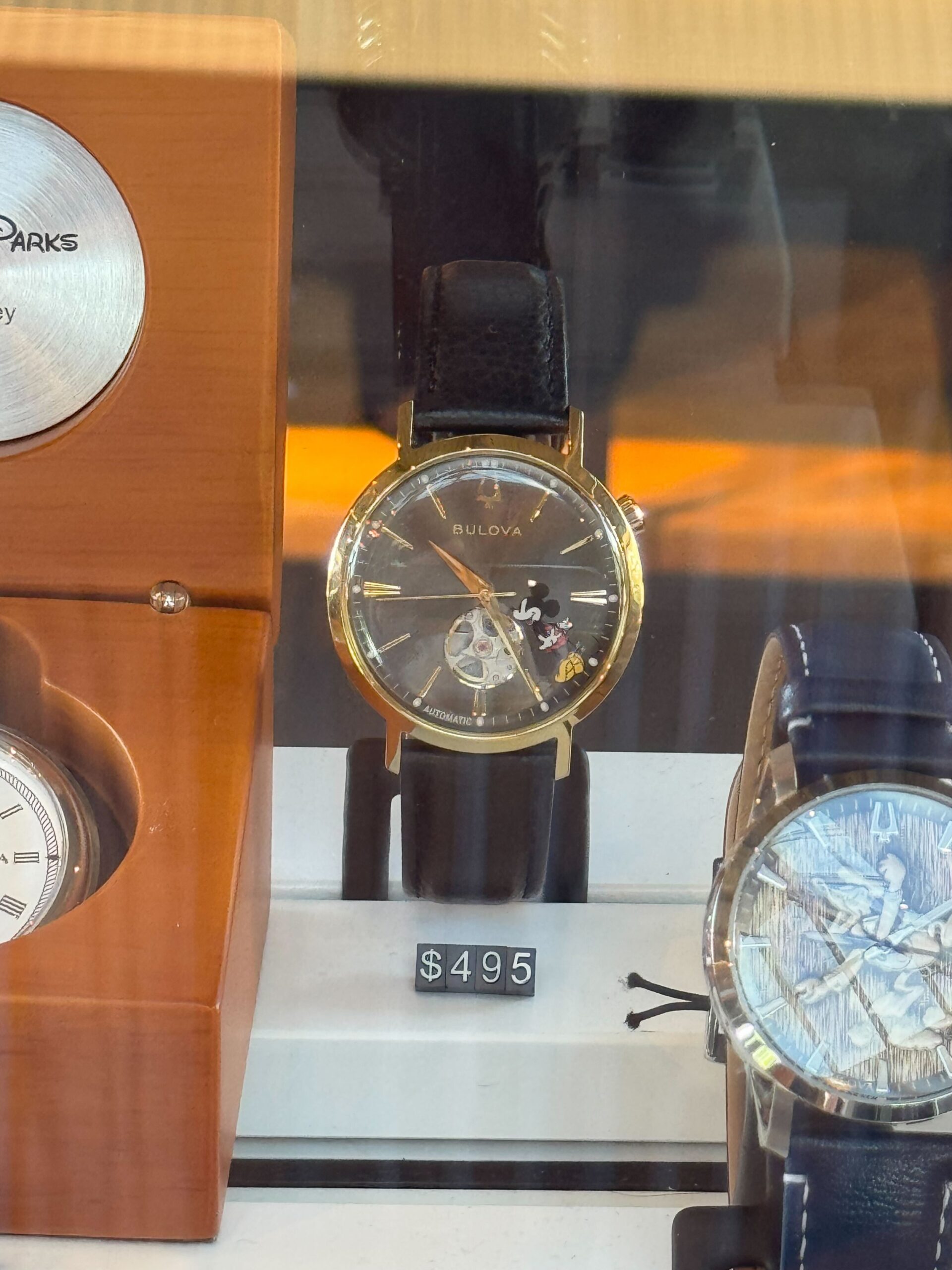 The Mickey watch by Bulova features a gold-tone round case paired with a black leather strap. The design of the black dial shows Mickey standing on his tip toes between the 4 and 5 numerals. There are gold-tone details on the strap and crown, as well as the numbers on the face.
