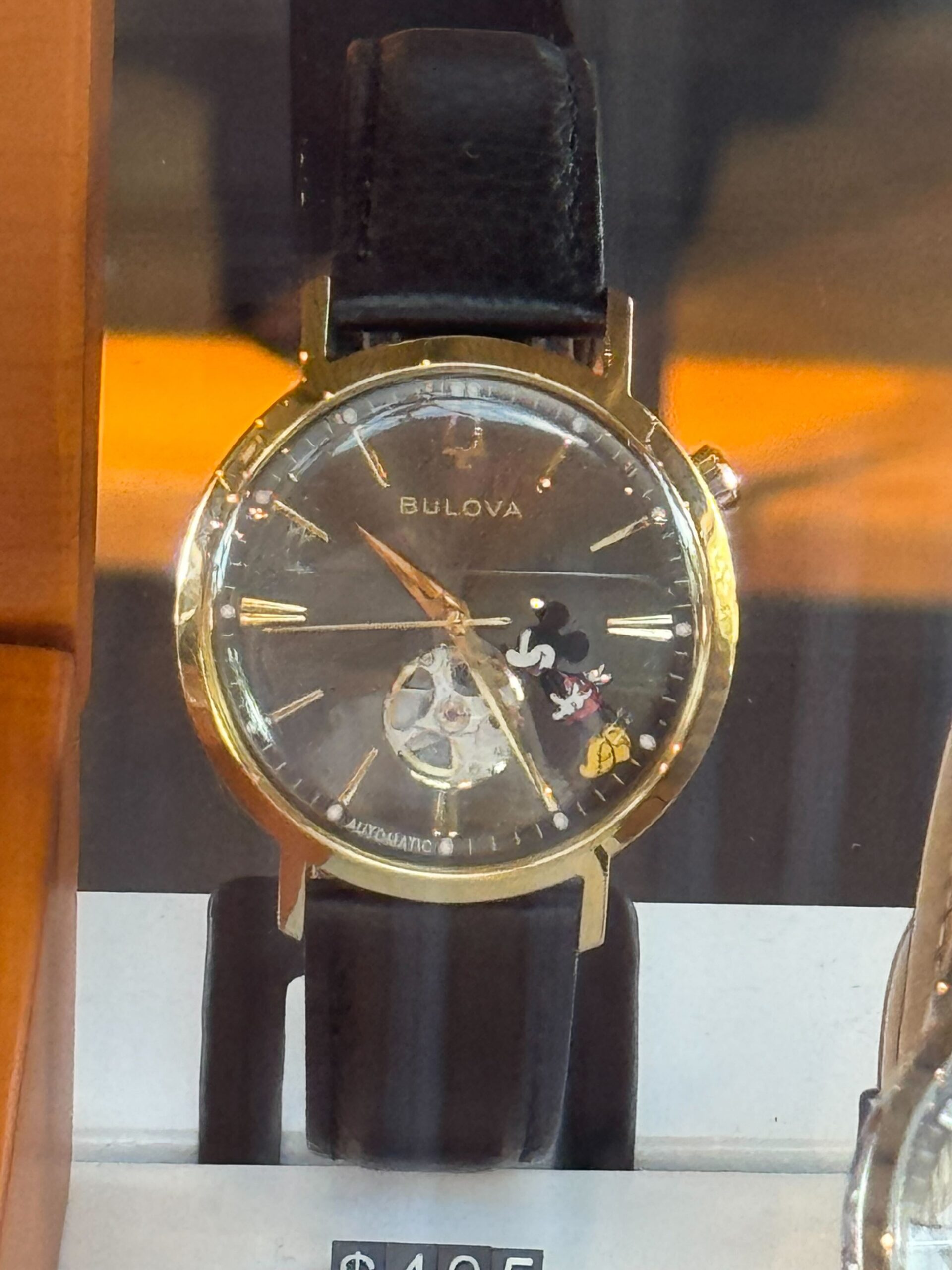 The Mickey watch by Bulova features a gold-tone round case paired with a black leather strap. The design of the black dial shows Mickey standing on his tip toes between the 4 and 5 numerals. There are gold-tone details on the strap and crown, as well as the numbers on the face.