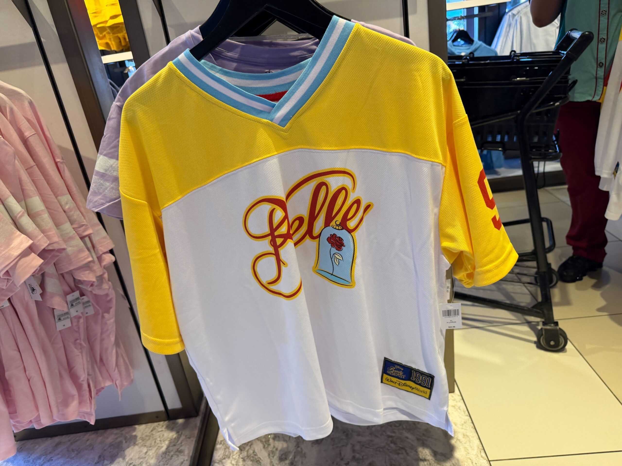 This Beauty and the Beast jersey is white and yellow with blue and white striping at the collar. The front features “Belle” in a red script outlined in a yellow tone, with a small graphic of the rose below.