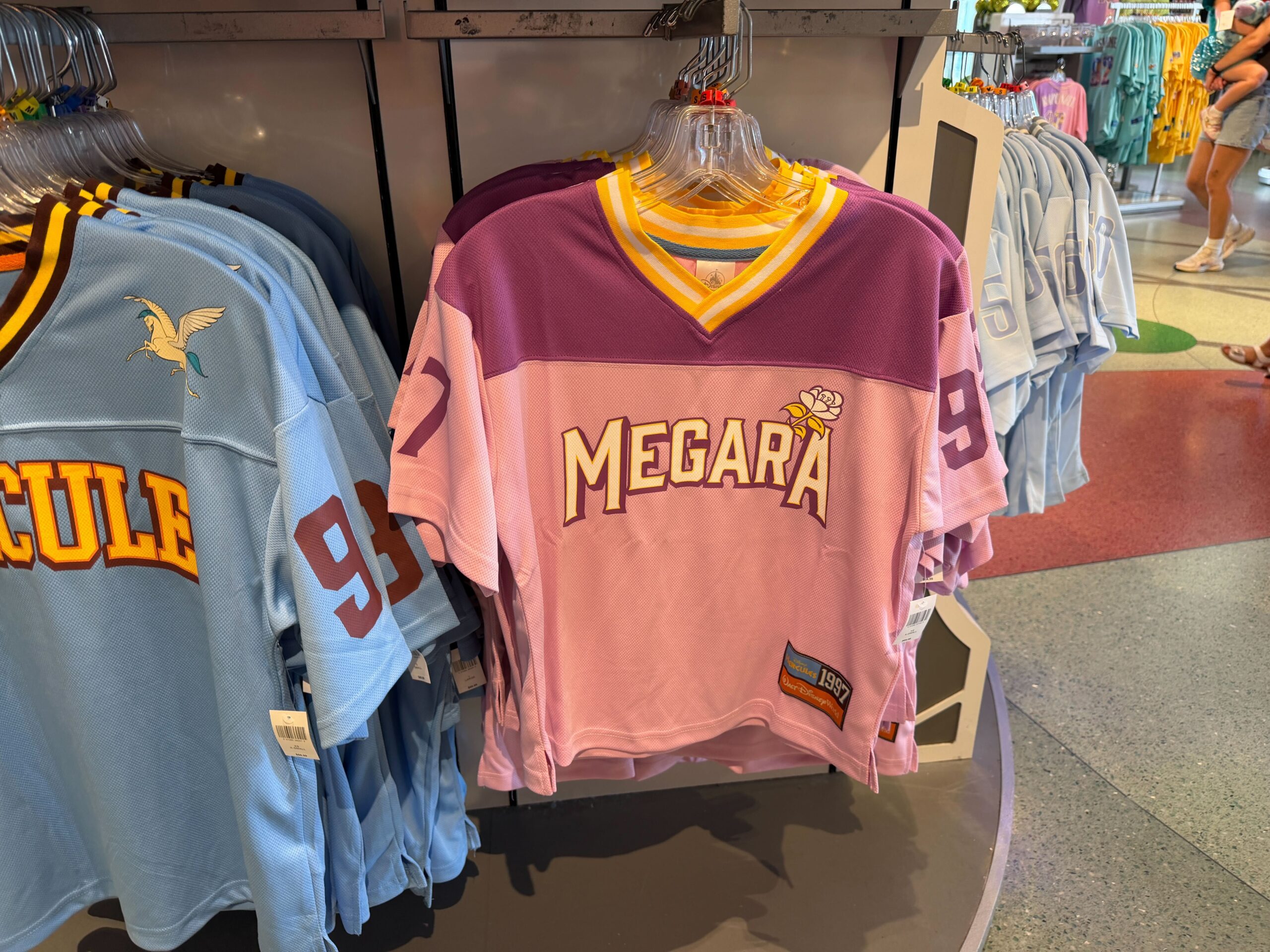 The jersey is a light pink and purple with white and gold striping at the V-neck, and matching athletic styling. “Megara” arcs across the chest in white collegiate lettering in gold with a burgundy shadow. A flower sits above the "A."