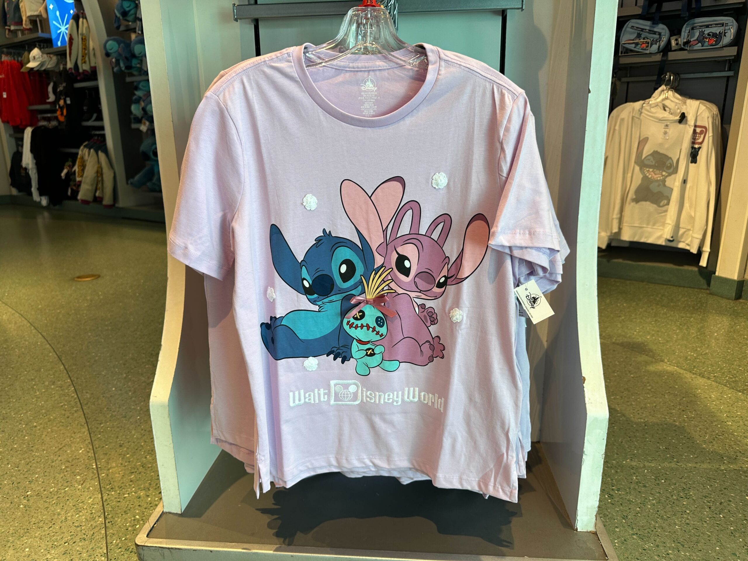 This pink tee has an image of Stitch and Angel propped up against each other with Scrump in the middle. White flowers are around them, and the Disney World wordmark is at the bottom.