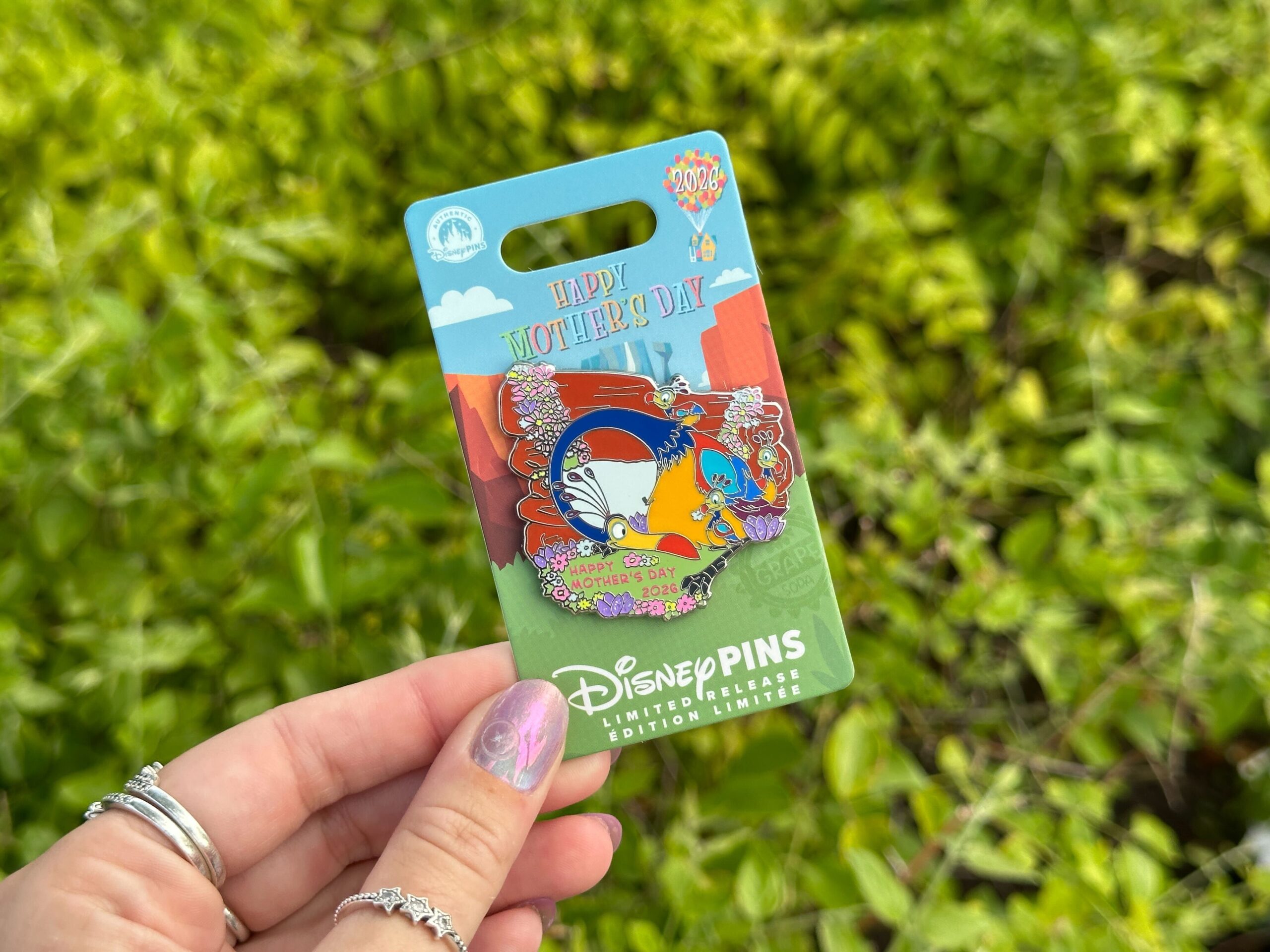 This year's Mother's Day pin features Kevin and her babies from Up. The bird is depicted by a rocky archway with pastel flowers growing around. She has her neck curled down to look at her trio of chicks.