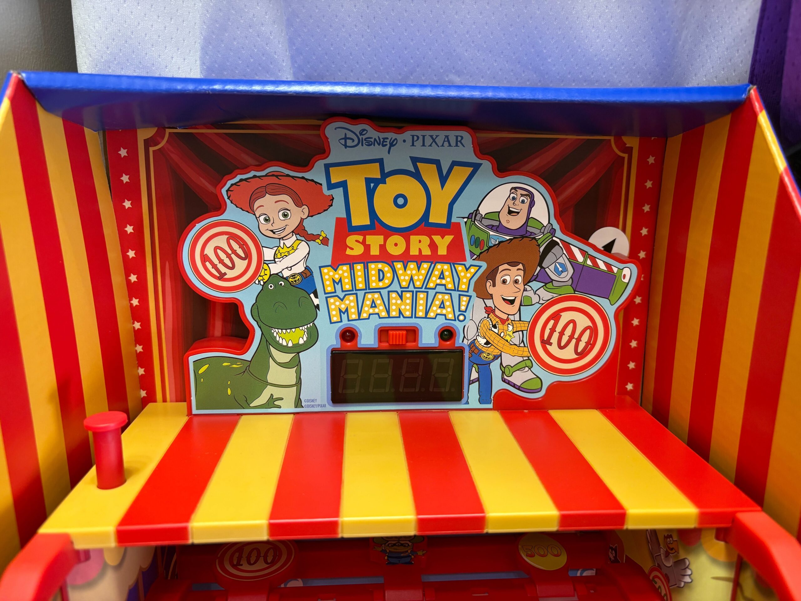 This plastic toy is a small version of a carnival game booth. It has a built-in shooter and shelves. A marquee features the "Toy Story Midway Mania!" logo and images of Jessie, Rex, Woody, and Buzz.
