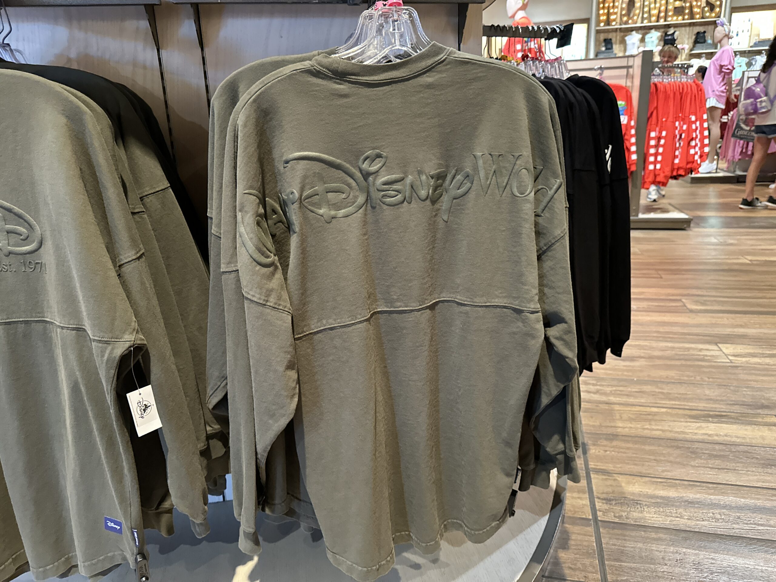 This is a muted army green color. It has a cursive "D" logo on the left chest with "Est. 1971" — the year Walt Disney World opened. "Walt Disney World" is across the back. The text is green puffy ink.