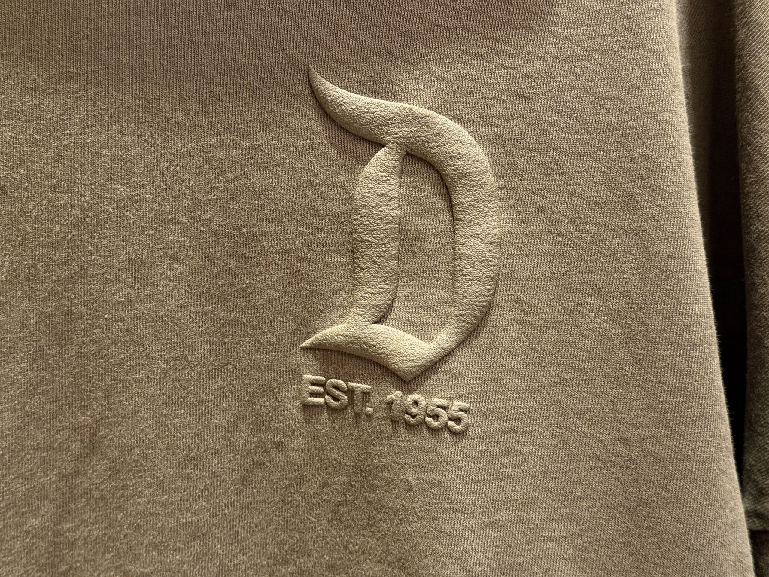 This is a muted army green color. It has the gothic Disneyland "D" logo on the left chest with "Est. 1955" — the year Disneyland opened. The text is puffy ink the same color as the fabric.