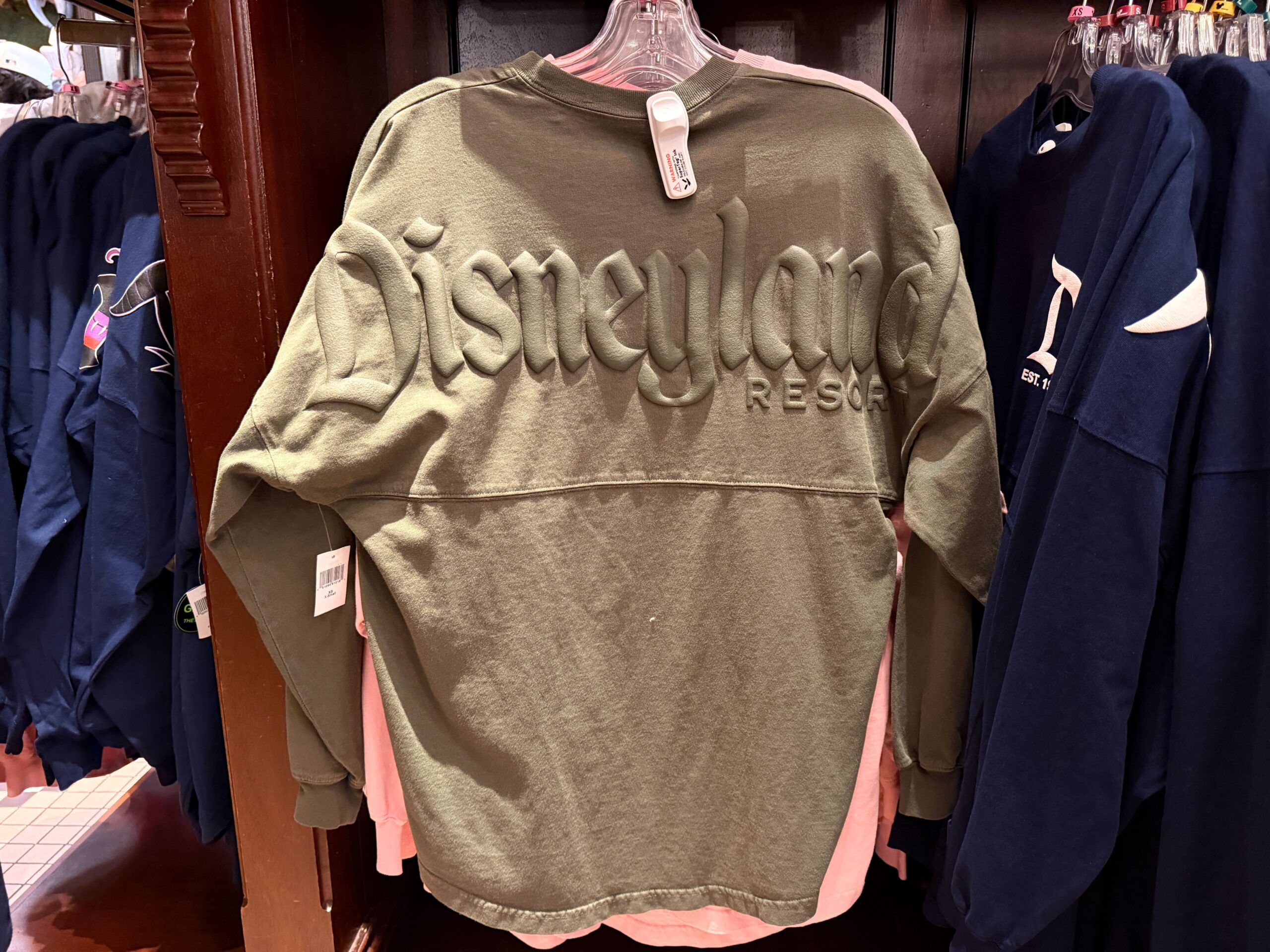 This is a muted army green color. It has the gothic Disneyland "D" logo on the left chest with "Est. 1955" — the year Disneyland opened. The text is puffy ink the same color as the fabric.