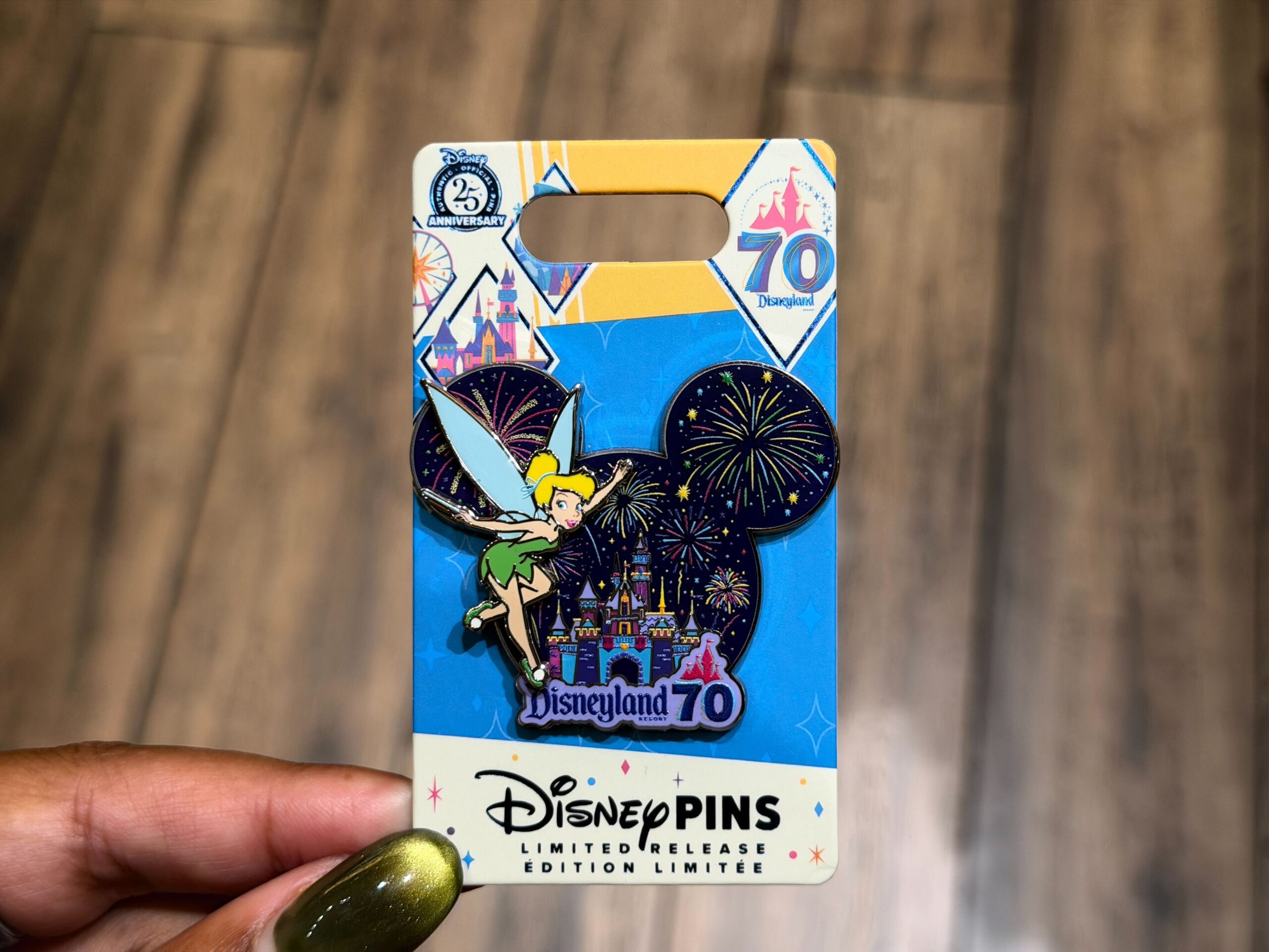 This pin is in the shape of Mickey's head and also has fireworks in a night sky. Sleeping Beauty Castle in corresponding colors is beneath the fireworks, and Tinker Bell in a simplistic design is flying to the left in the foreground. The Disneyland 70 logo is at the bottom.