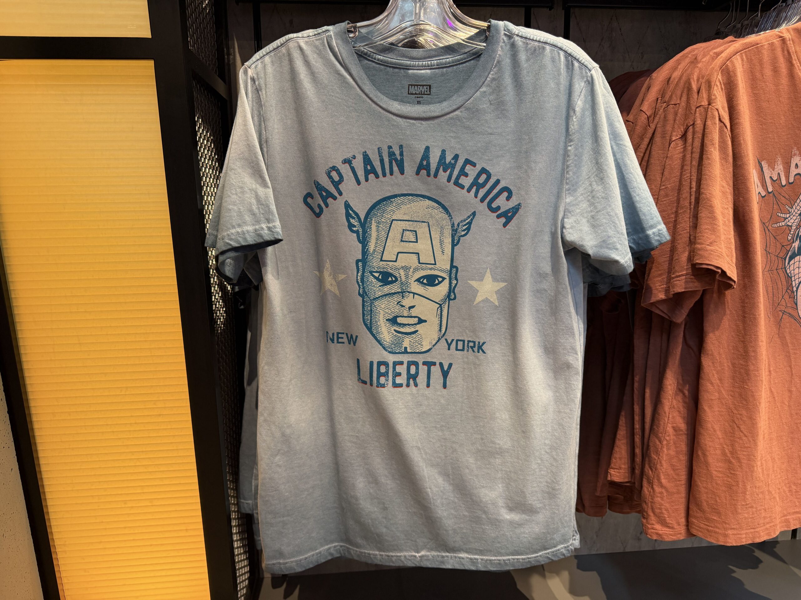 Captain America's face with his "A" mask is pictured on the center of this pale blue ombré tee. "Captain America - New York - Liberty" is in faded dark blue lettering with white stars.